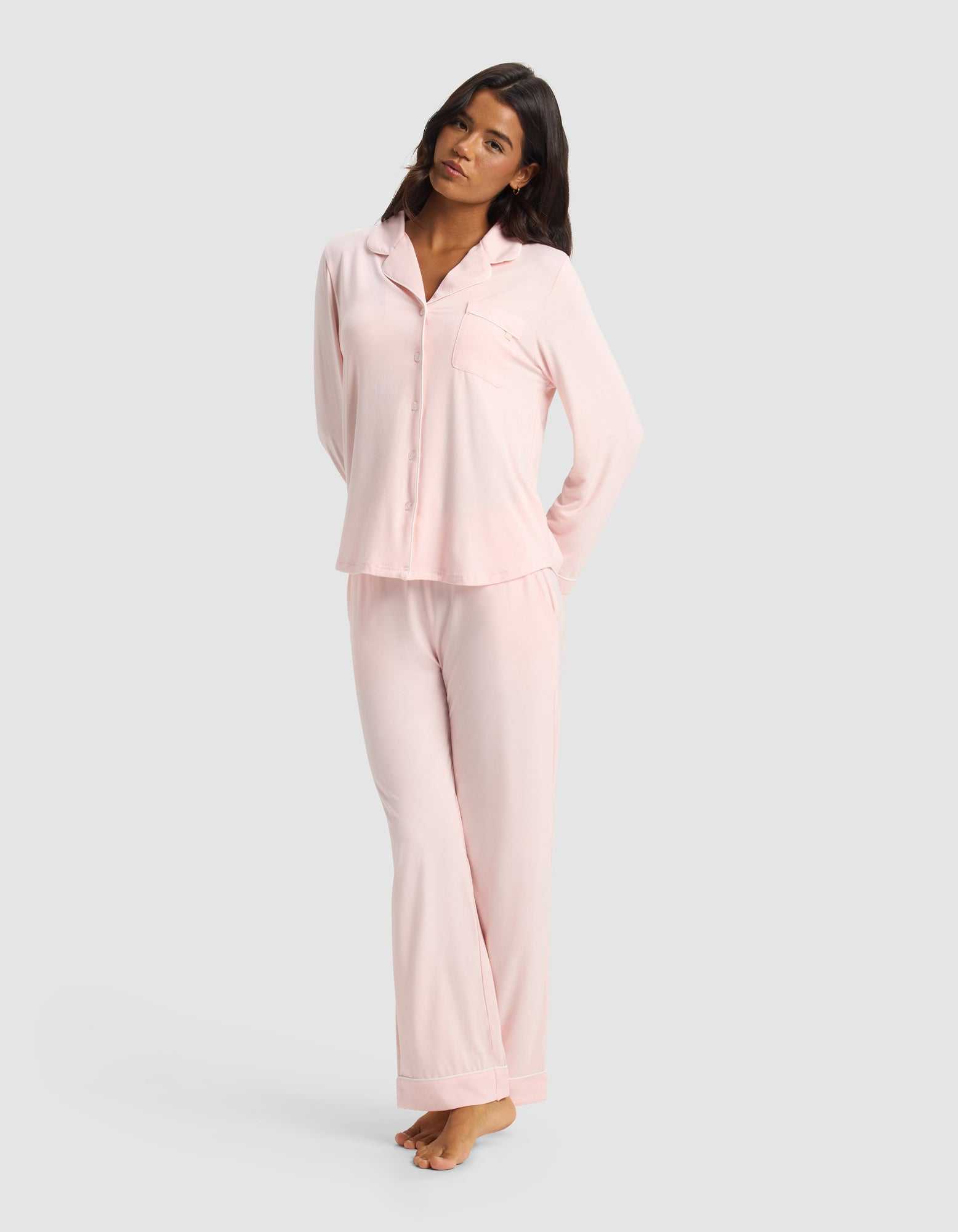 A woman with long dark hair stands barefoot against a light gray background, wearing Cozy Earth’s Women's Bamboo Stretch-Knit Long Sleeve Pajama Set—light pink, with a button-up top and matching pants. Her arms are relaxed behind her back.|Color:Blossom