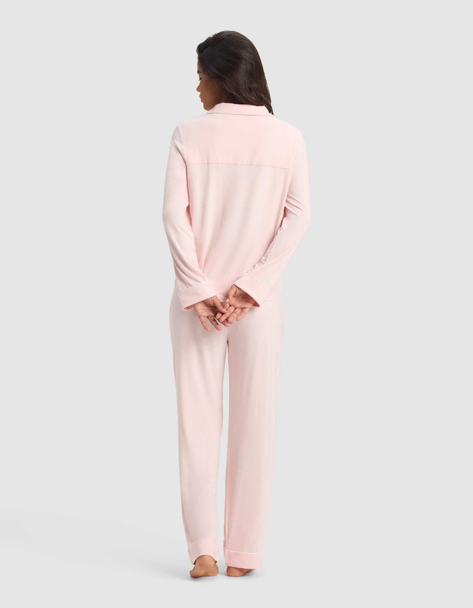 A woman stands barefoot facing away in the Cozy Earth Women's Bamboo Stretch-Knit Long Sleeve Pajama Set in light pink. Her dark hair is loose, hands clasped behind her back, against a plain light gray background.|Color:Blossom