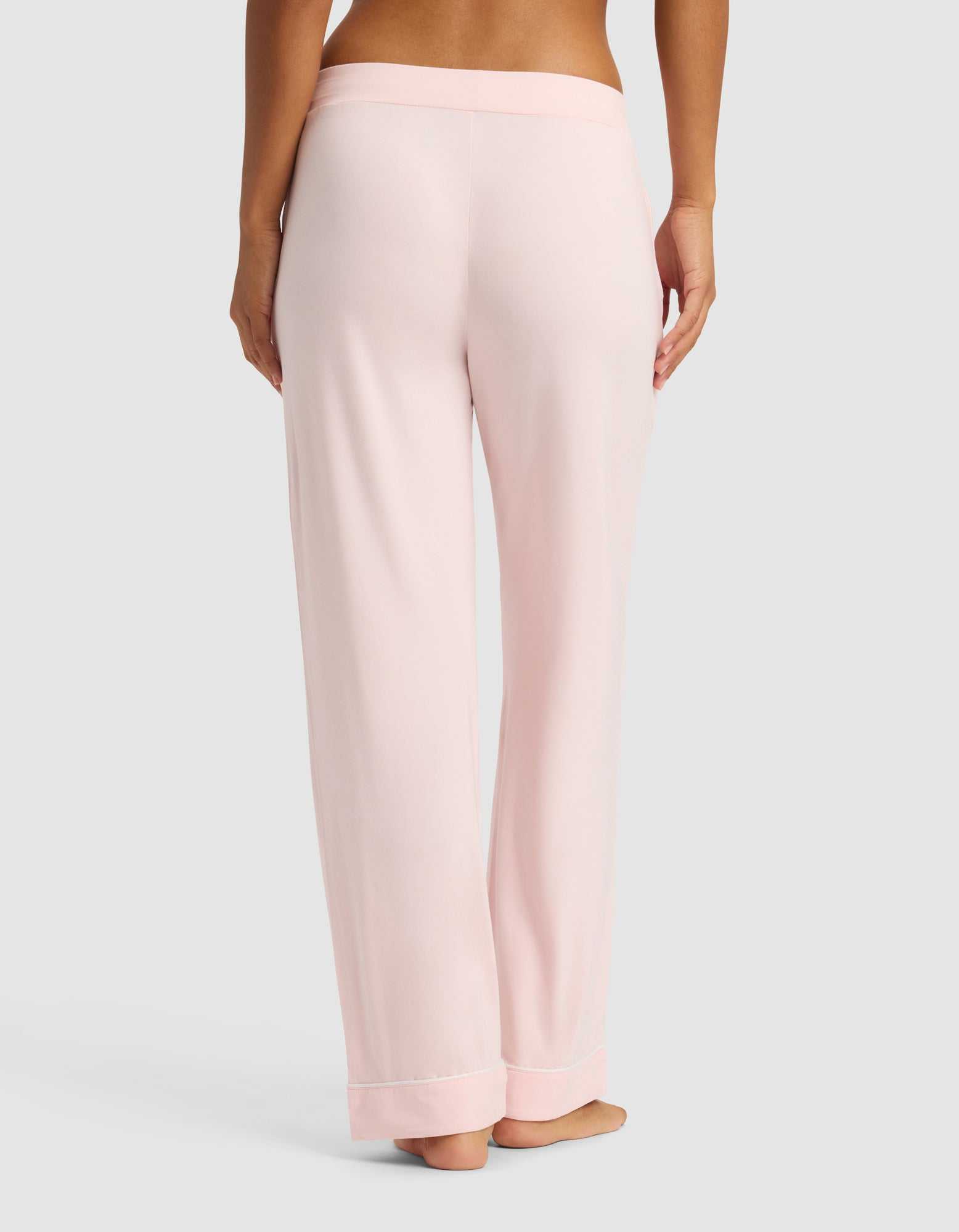 A woman is shown from the waist down wearing Cozy Earth Women's Bamboo Stretch-Knit Long Sleeve Pajama Set pants in light pink, standing barefoot against a plain gray background.|Color:Blossom