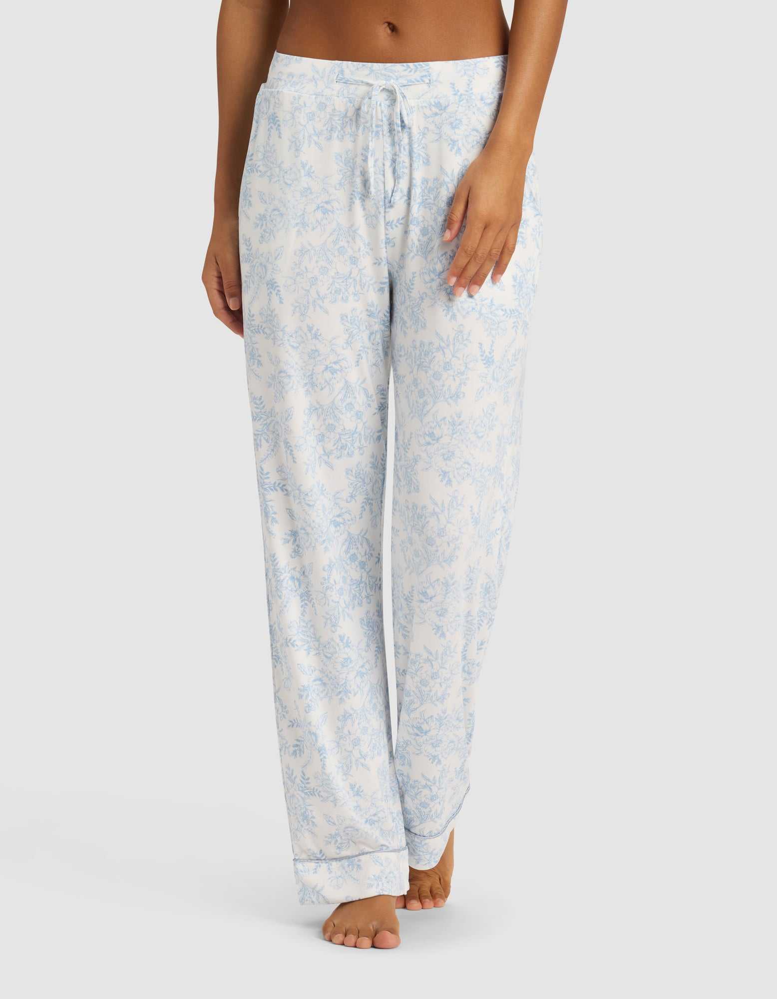 A woman wears Cozy Earth's Women's Bamboo Stretch-Knit Long Sleeve Pajama Set in light blue and white floral, featuring a drawstring waist, standing barefoot against a plain white background.|Color:Sky Toile