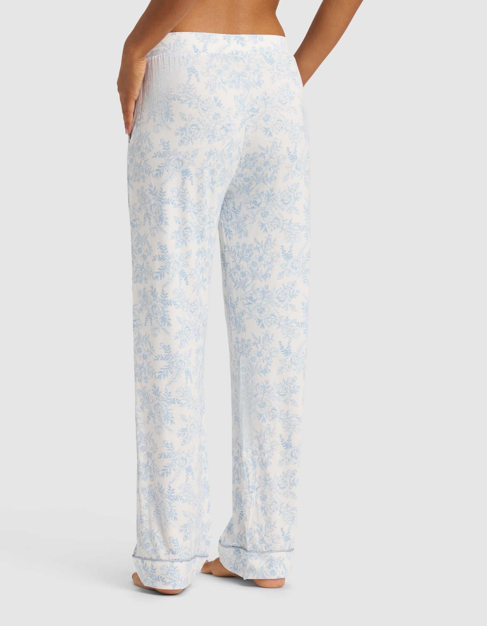 A person stands barefoot facing away, wearing Cozy Earth's Women's Bamboo Stretch-Knit Long Sleeve Pajama Set in white with a light blue floral pattern. Their right hand is on their hip against a plain light gray background.|Color:Sky Toile