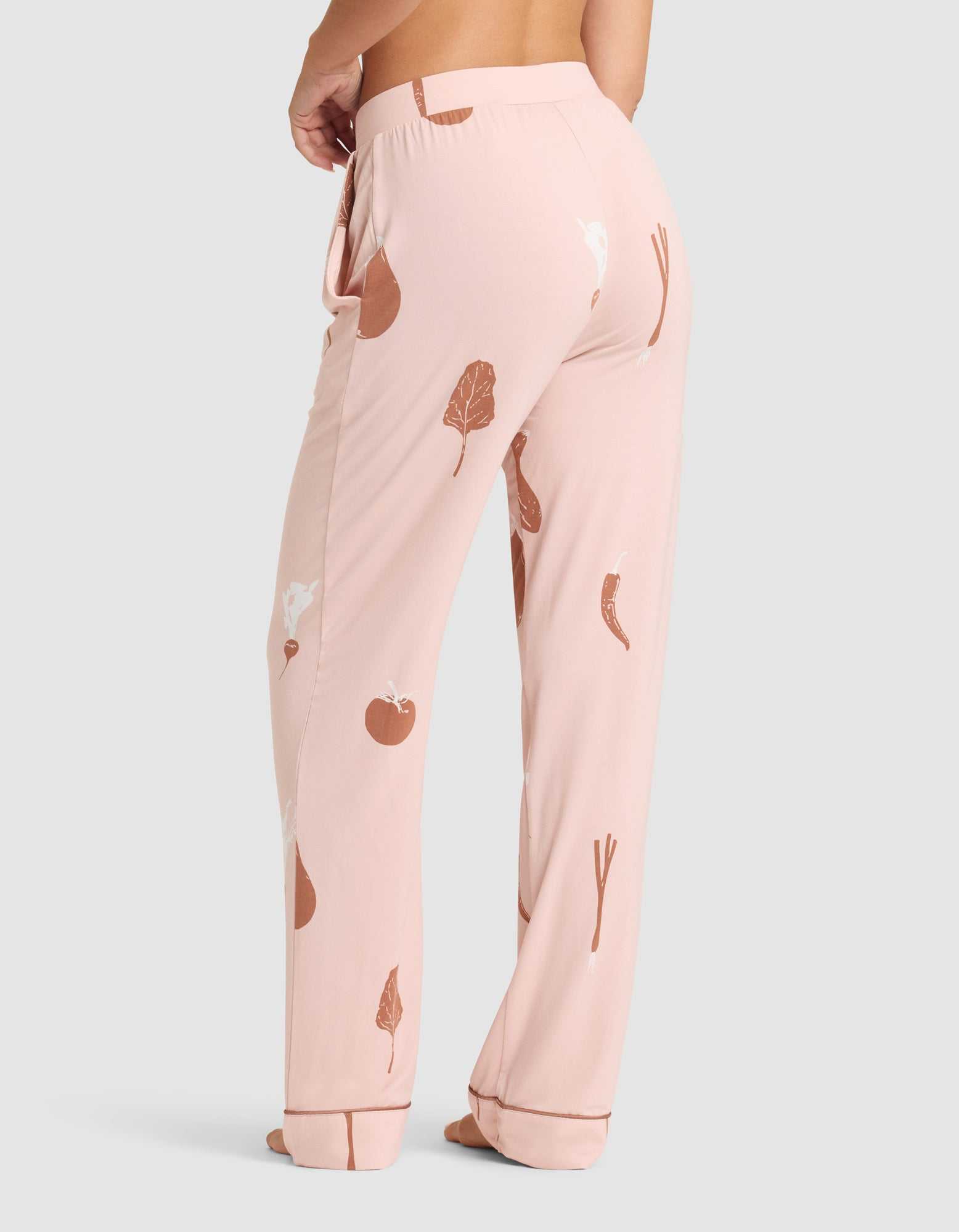 A person is barefoot wearing HIDE Women's Bamboo Stretch-Knit Pajama Pant in light pink with brown and white vegetable prints, shown from the lower back and legs against a plain light gray background. |Color:Veggies in Dusty Rose