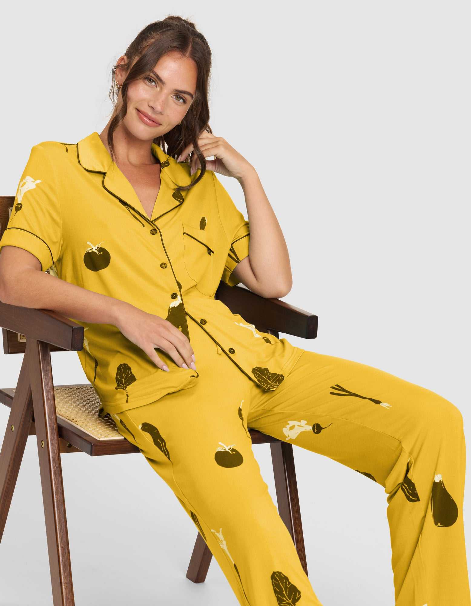 A woman with long brown hair smiles softly as she sits on a wooden chair, wearing Cozy Earth’s Women's Bamboo Stretch-Knit Short Sleeve & Pant Pajama Set (Veggie), featuring a yellow design with dark vegetable illustrations. |Color:Veggies in Mango
