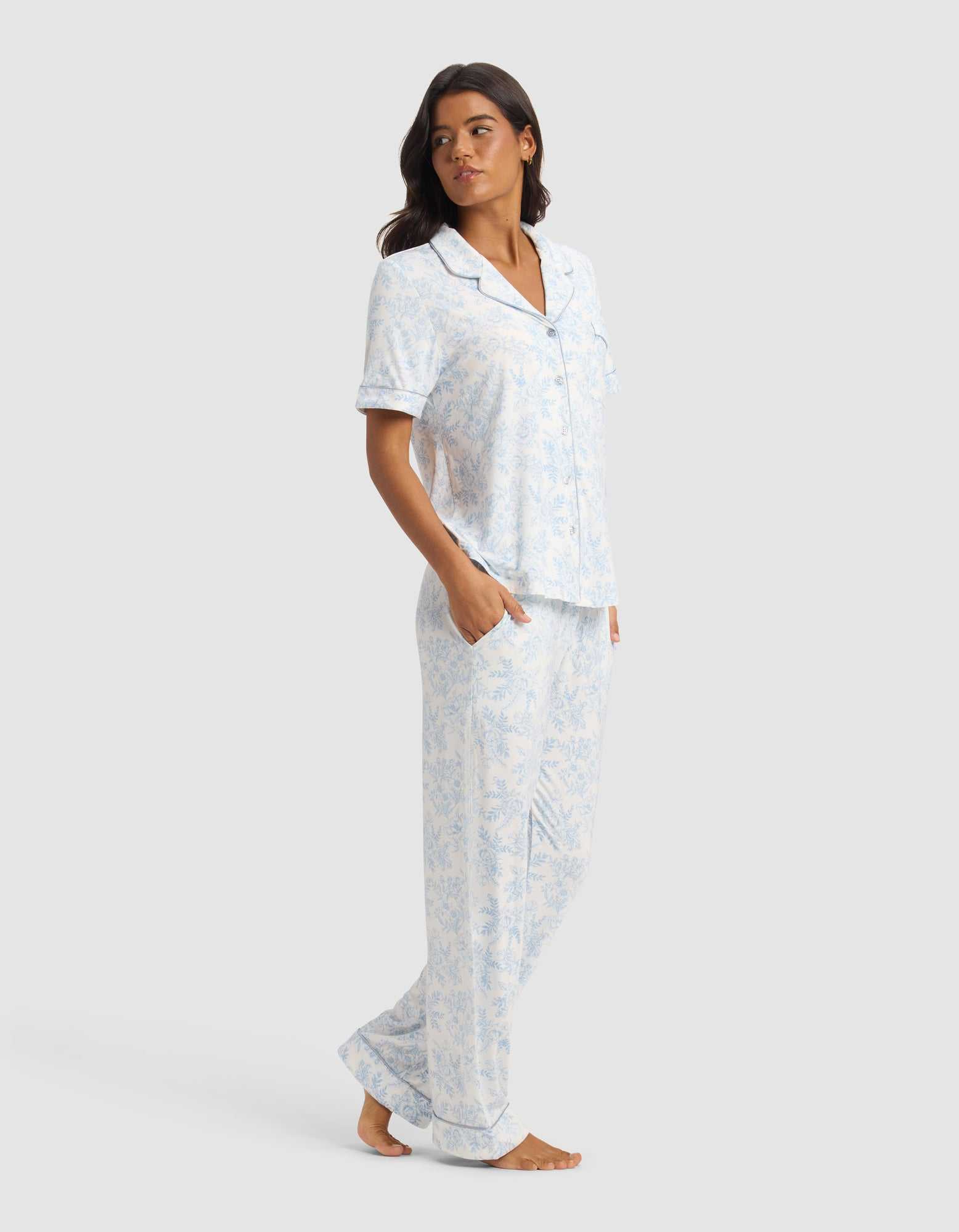 A woman stands barefoot against a plain background, wearing the Cozy Earth Women's Bamboo Stretch-Knit Short Sleeve & Pant Pajama Set in light blue and white. The set features a collared, button-up top with chest pocket and matching pants. She looks to the side with hands in her pockets.|Color:Sky Toile