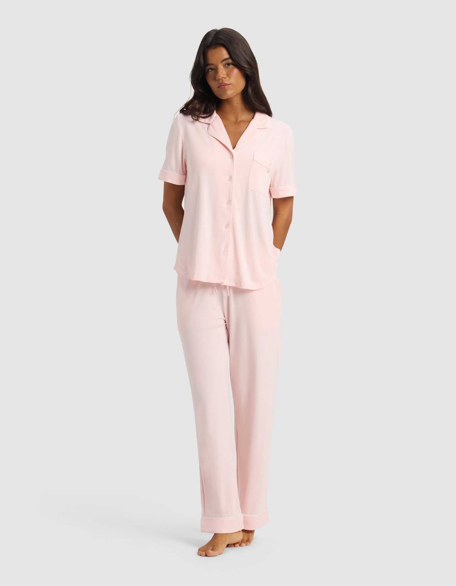 A woman stands barefoot against a plain light gray background, wearing the Cozy Earth Women's Bamboo Stretch-Knit Short Sleeve & Pant Pajama Set in Blossom, with her arms relaxed behind her back.|Color:Blossom