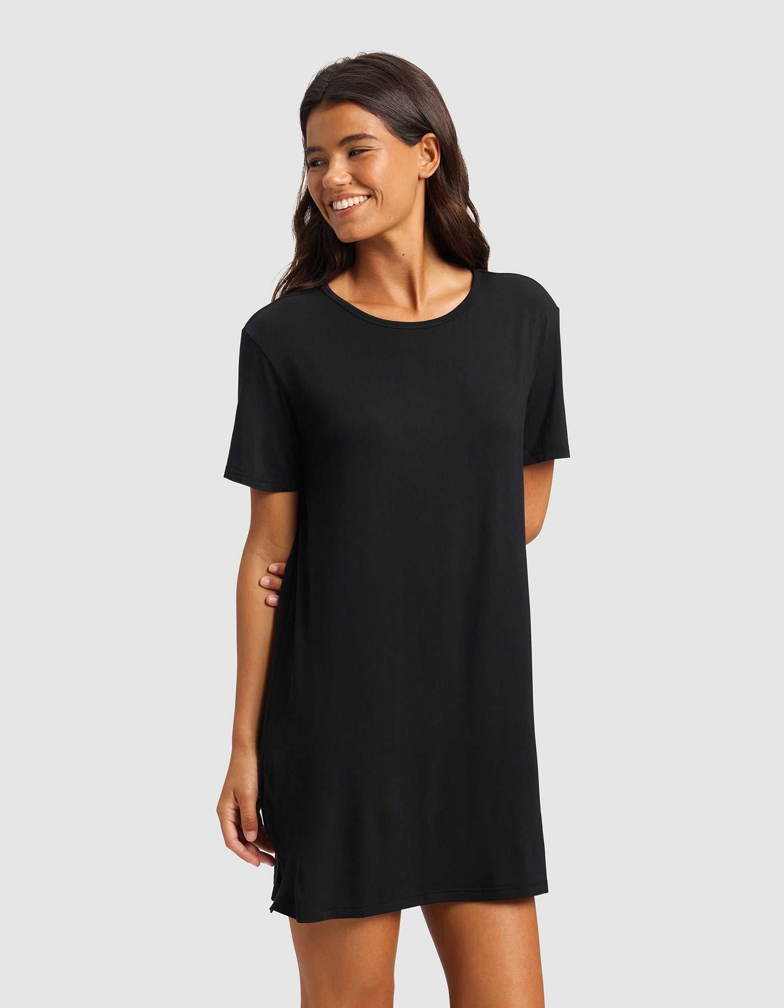 A woman with long dark hair smiles while wearing the Cozy Earth Women's Bamboo Stretch-Knit Sleep Dress, a simple black short-sleeved dress that falls above the knees, standing against a plain light gray background.|Color:Black
