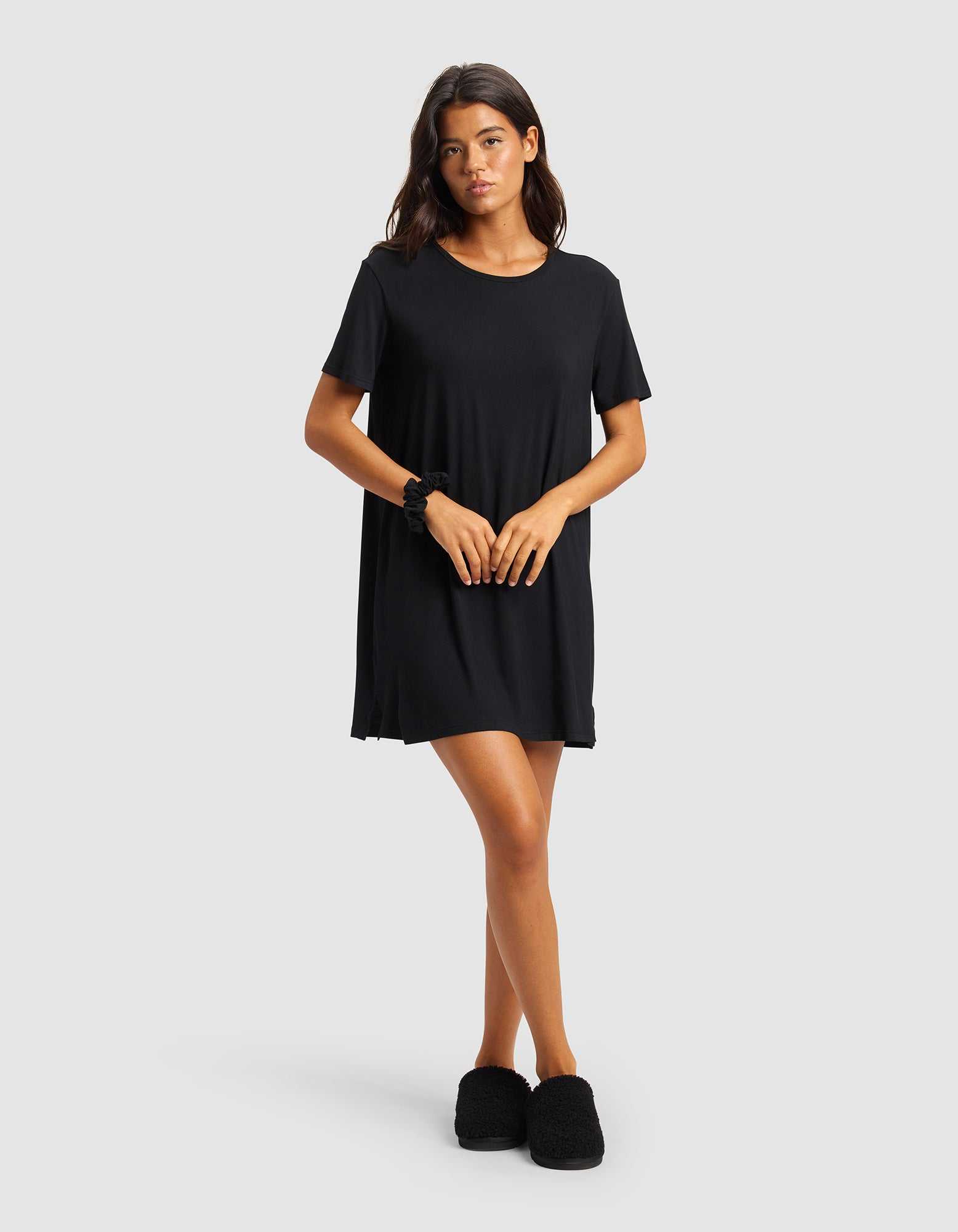 A woman stands against a plain light gray background, wearing Cozy Earth's Women's Bamboo Stretch-Knit Sleep Dress in black and matching slippers. She has long dark hair and her hands are gently clasped in front of her.|Color:Black