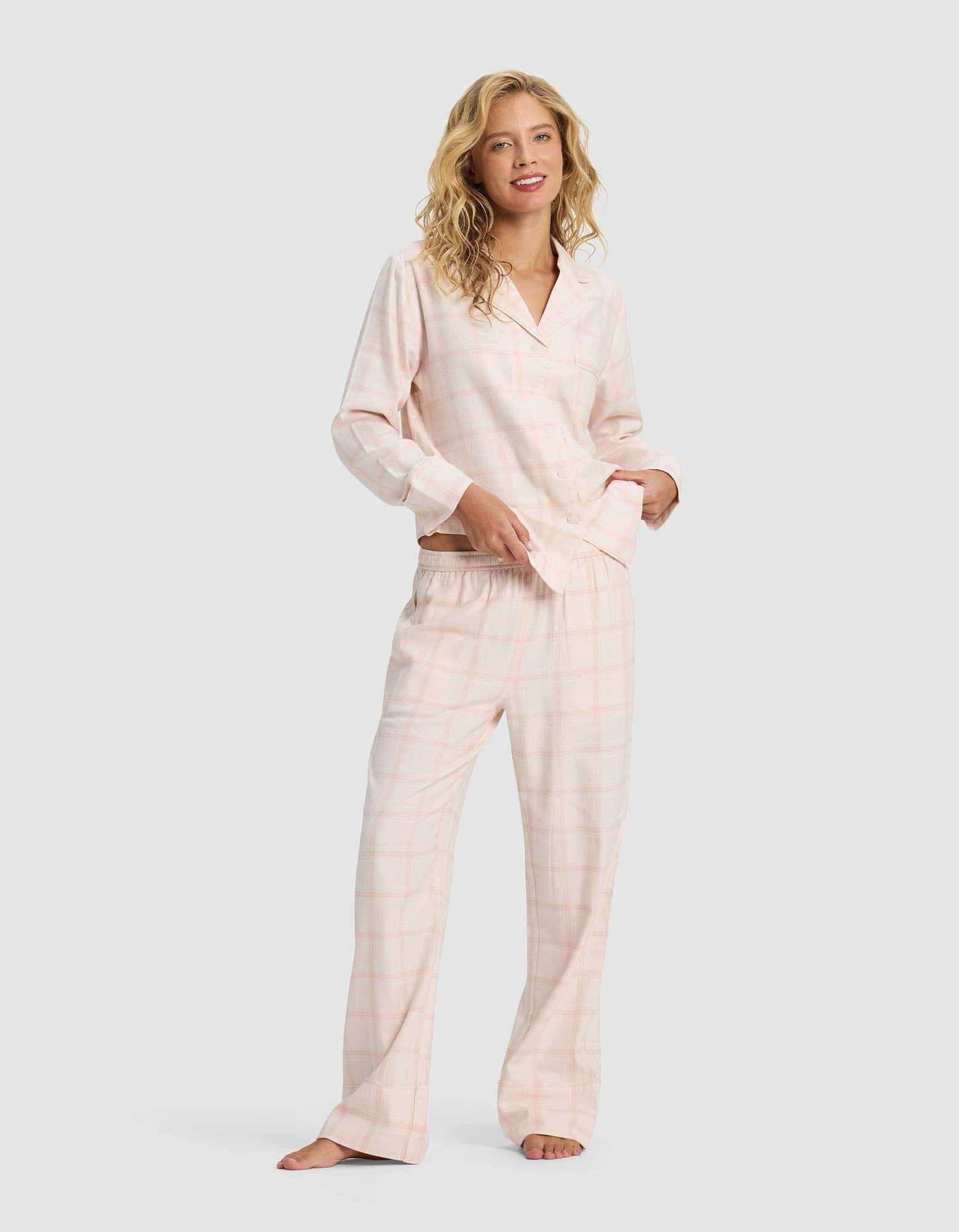 A woman with blonde hair wears Cozy Earth's Women's Farmhouse Pajama Pant in light pink plaid, standing barefoot and smiling against a plain light gray background.|Color:Quartz Plaid