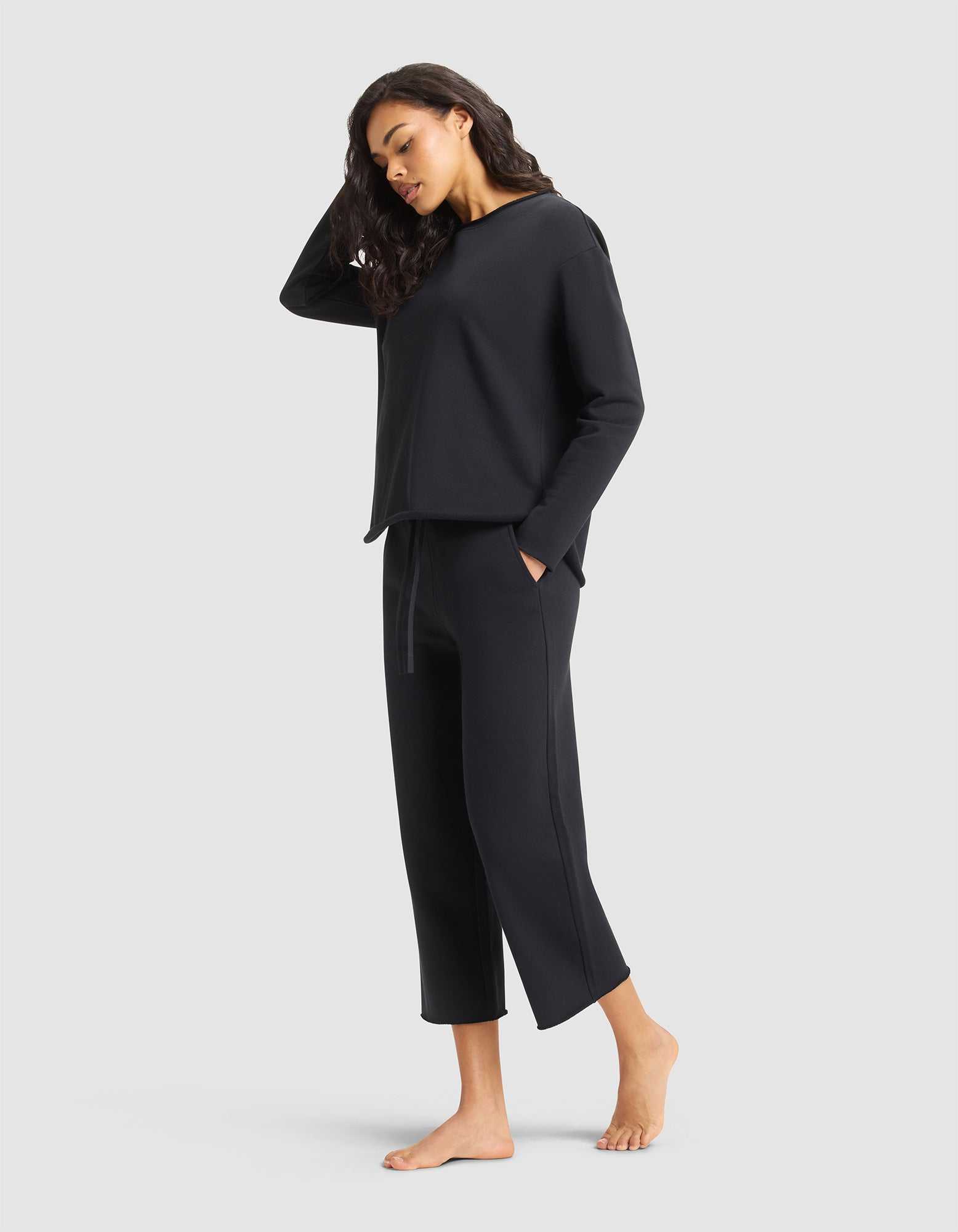 A woman with long dark hair stands barefoot against a light gray background, looking relaxed in the Cozy Earth Women's Chelsea Set—wearing a loose-fitting black top and matching cropped pants with her hands in her pockets. |Color:Jet Black