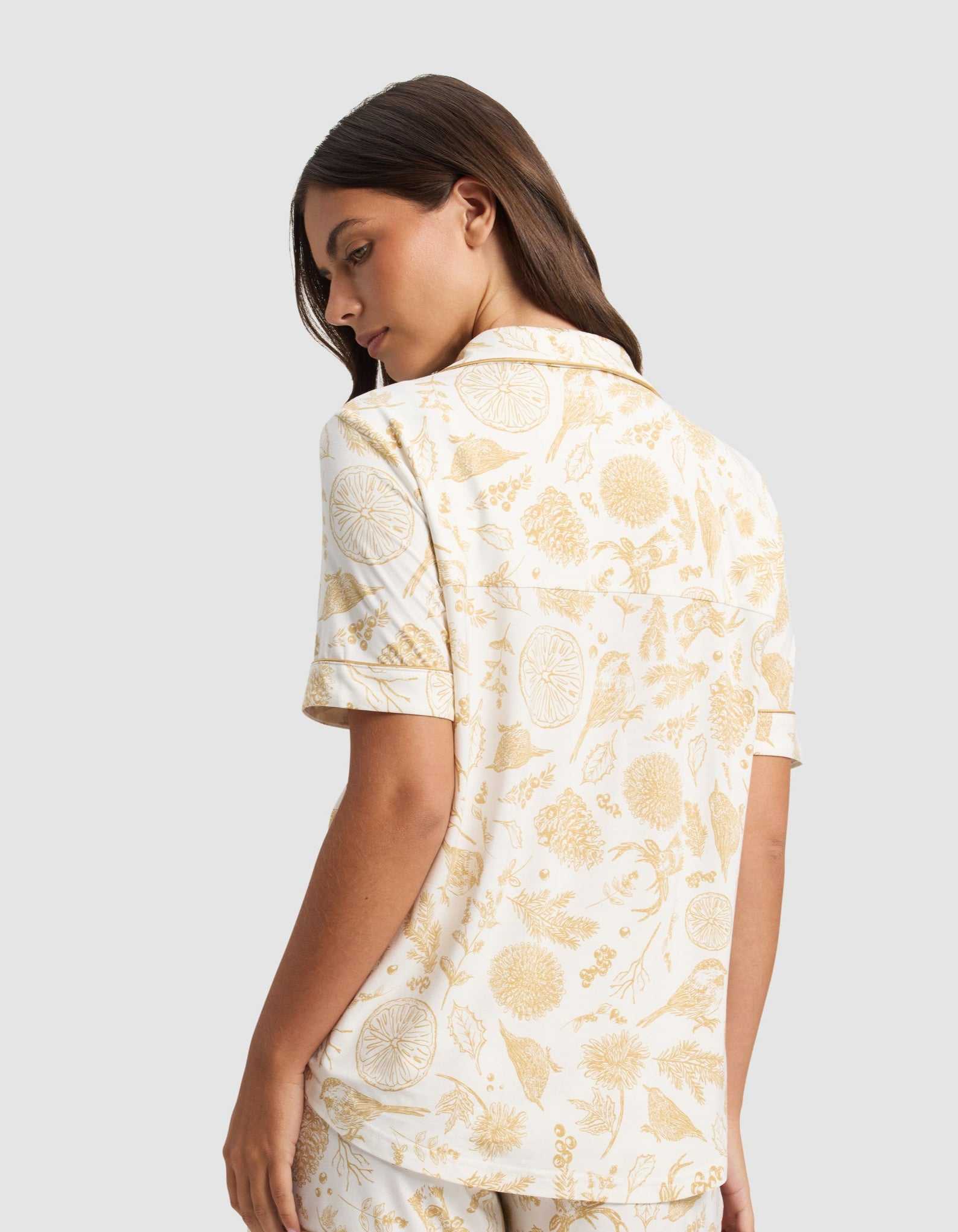 A woman with long brown hair stands turned to the side, wearing the Cozy Earth Women's Bamboo Stretch-Knit Short Sleeve Pajama Top featuring a yellow botanical and citrus fruit print on a white background.|Color:Winter Toile Dijon