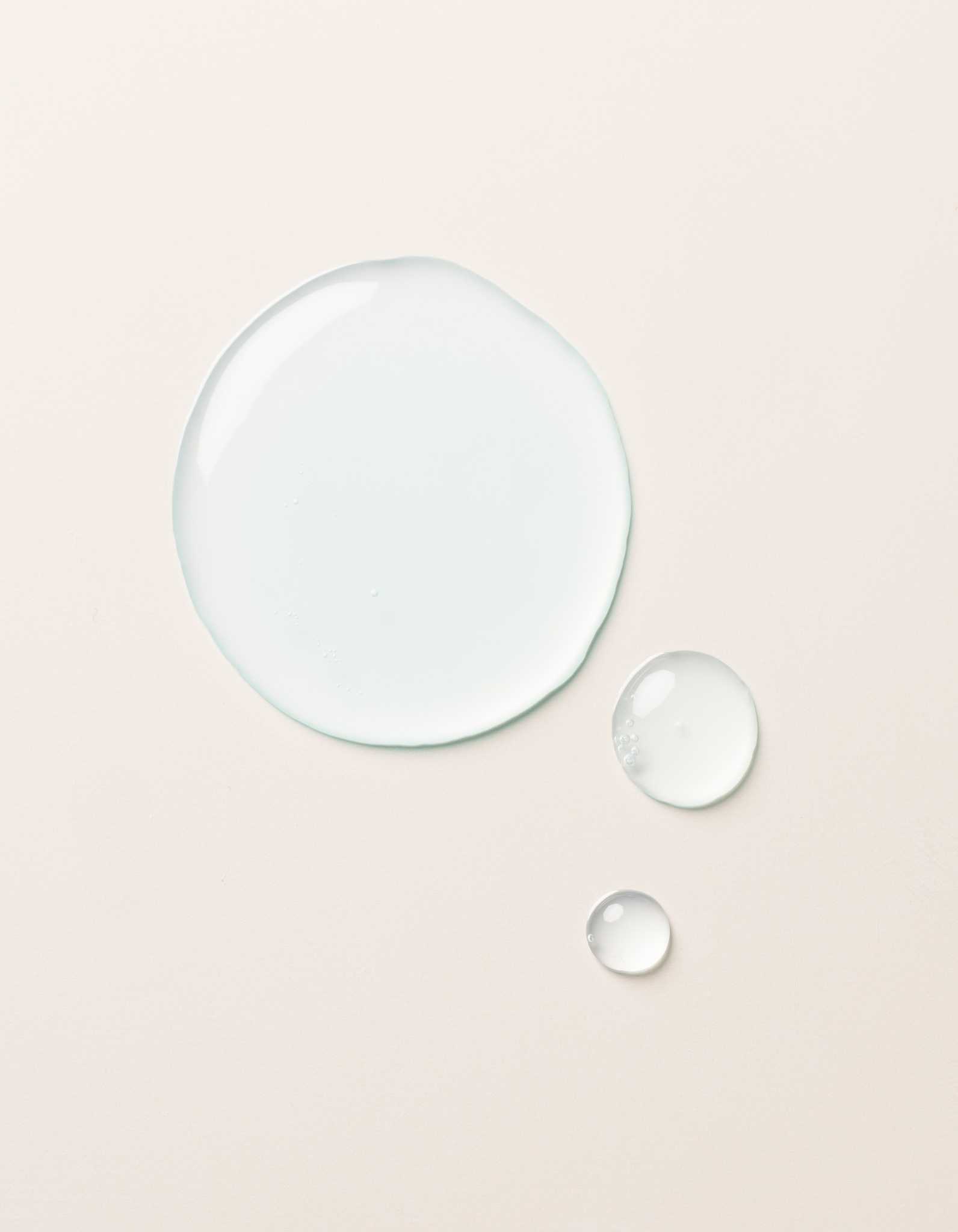 The Hydration Duo by Cozy Earth is shown as a large drop and two smaller drops of clear, light blue liquid on a smooth, light beige surface. The drops vary in size but all maintain a rounded shape, appearing glossy and slightly reflective.