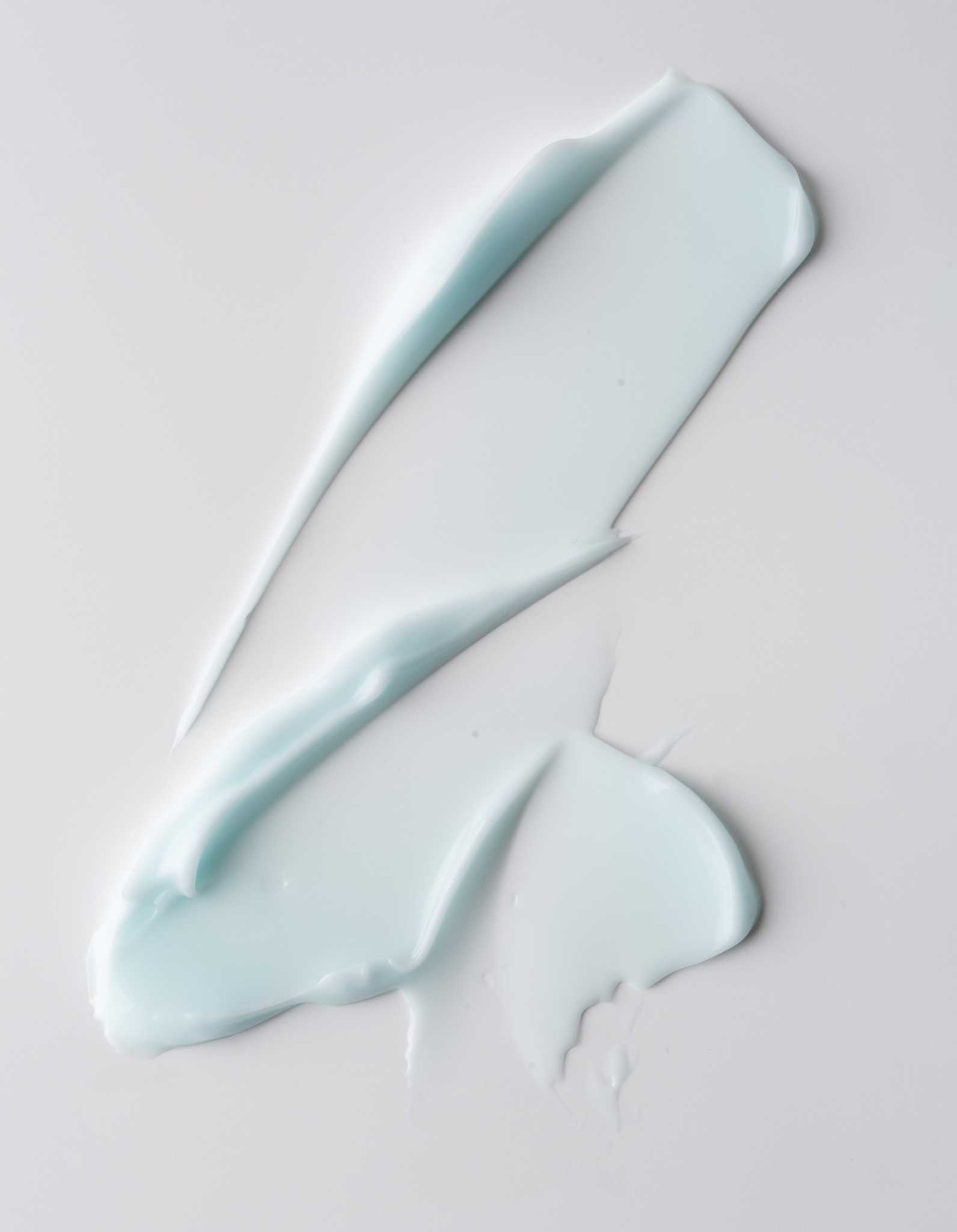 A smear of The Hydration Duo, a light blue, creamy substance from Cozy Earth, spread across a white surface. The texture appears smooth and glossy, indicating it could be a skincare product like cream, lotion, or gel.