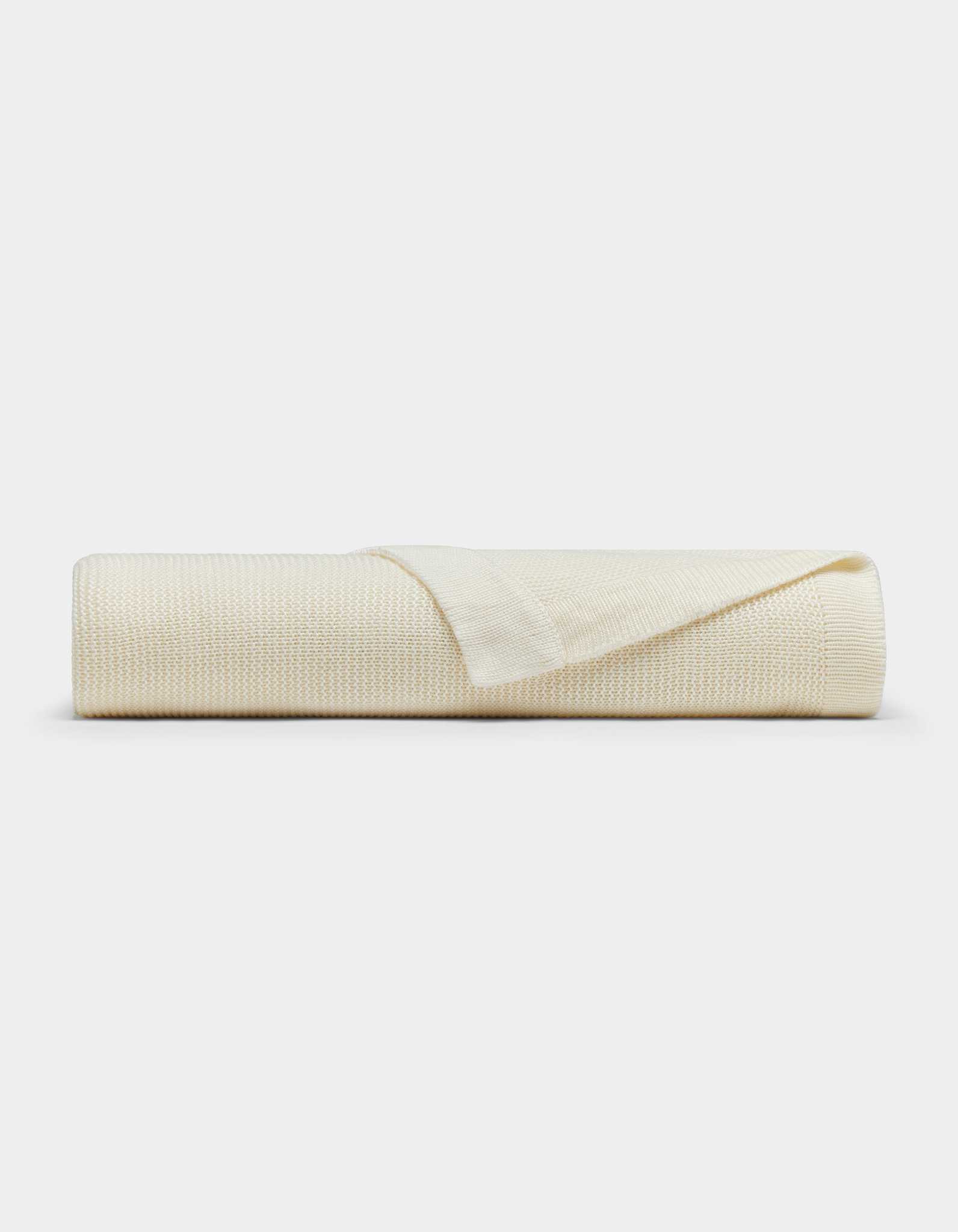 The Cozy Earth Cloud Knit Blanket by Cozy Earth is cream-colored with a textured pattern, neatly rolled and displayed on a white background. One edge is folded back, highlighting the blanket's thickness and texture.|Color:Ivory