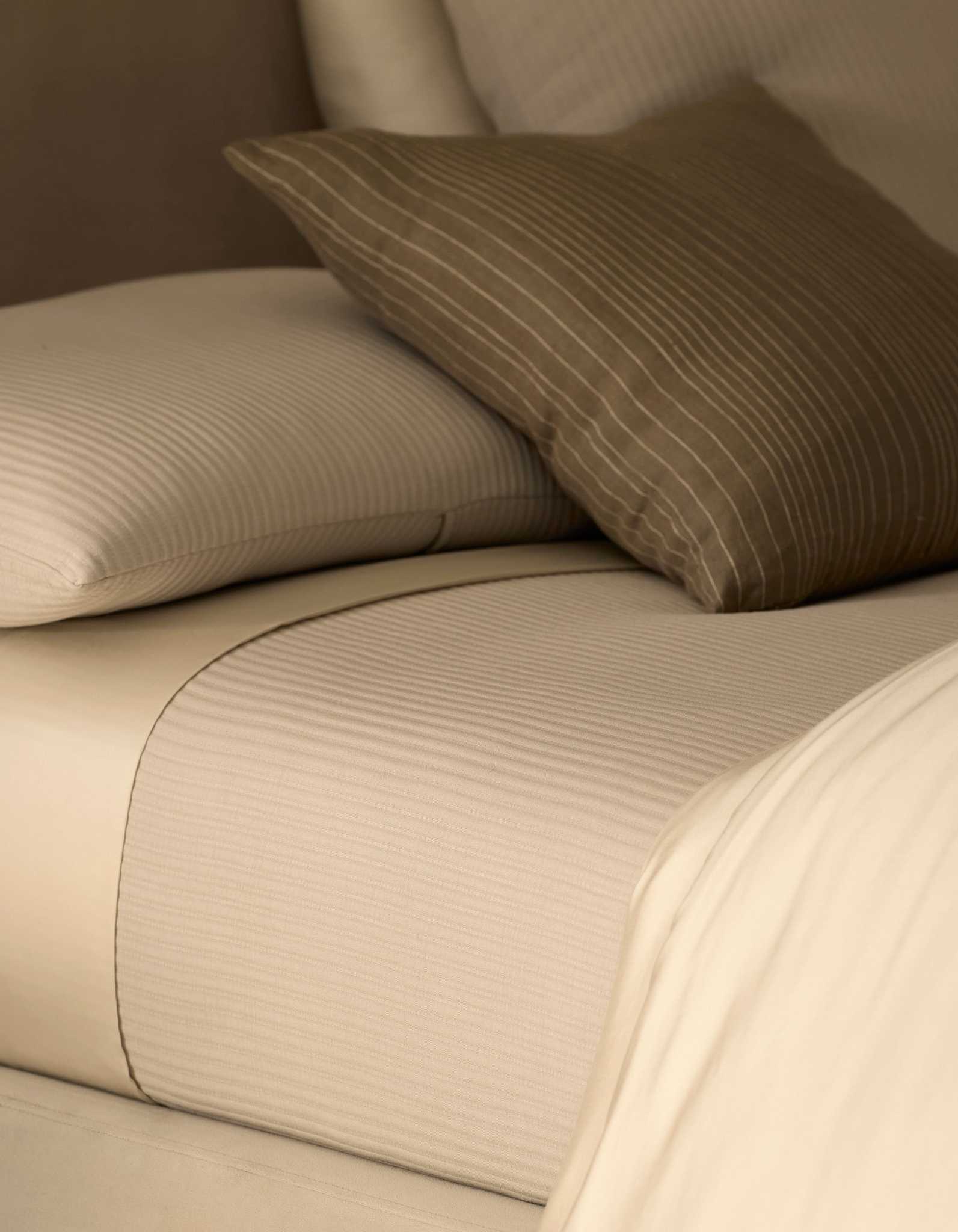 Close-up of a neatly made bed featuring the Cozy Earth Bamboo Coverlet in cream with subtle stripes, paired with a matching pillow and a brown accent pillow adorned with white pinstripes.|Color:Driftwood