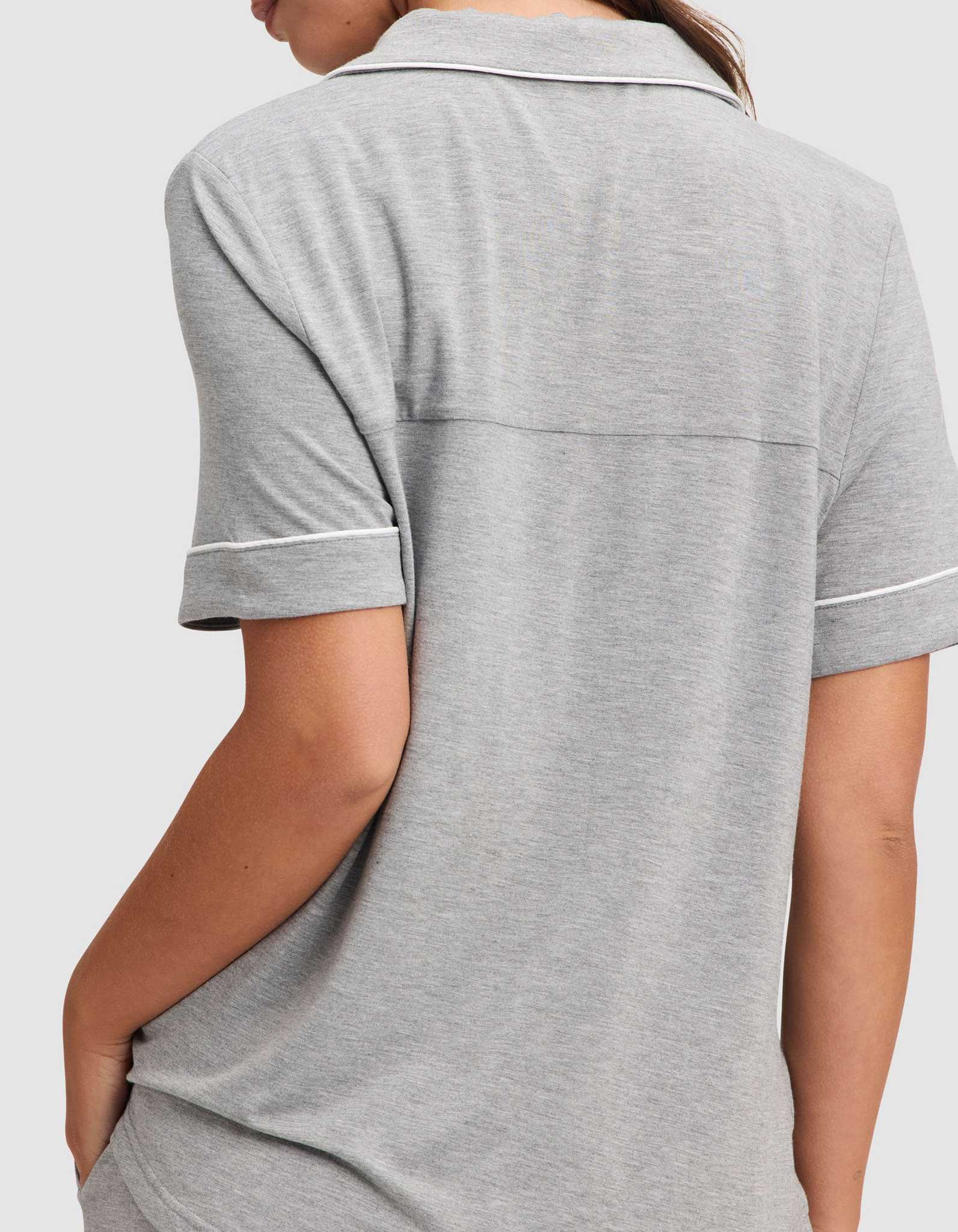A woman is shown from the back wearing the Cozy Earth Women's Bamboo Stretch-Knit Short Sleeve Pajama Set in light gray, featuring a collared top with white trim and short sleeves, with her left arm bent and hand in her pocket. |Color:Grey