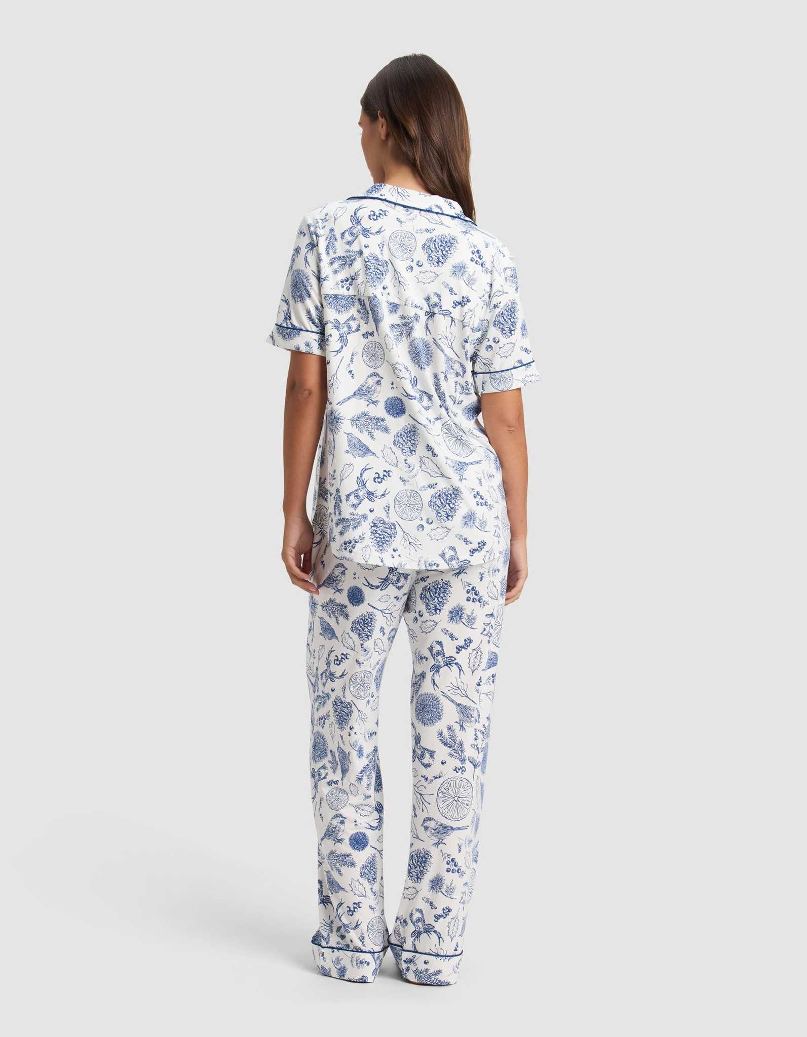 A woman with long brown hair faces away, wearing the Cozy Earth Women's Bamboo Stretch-Knit Short Sleeve Pajama Set in a blue and white botanical pattern with navy trim, set against a plain light gray background.|Color:Winter Toile Pacific