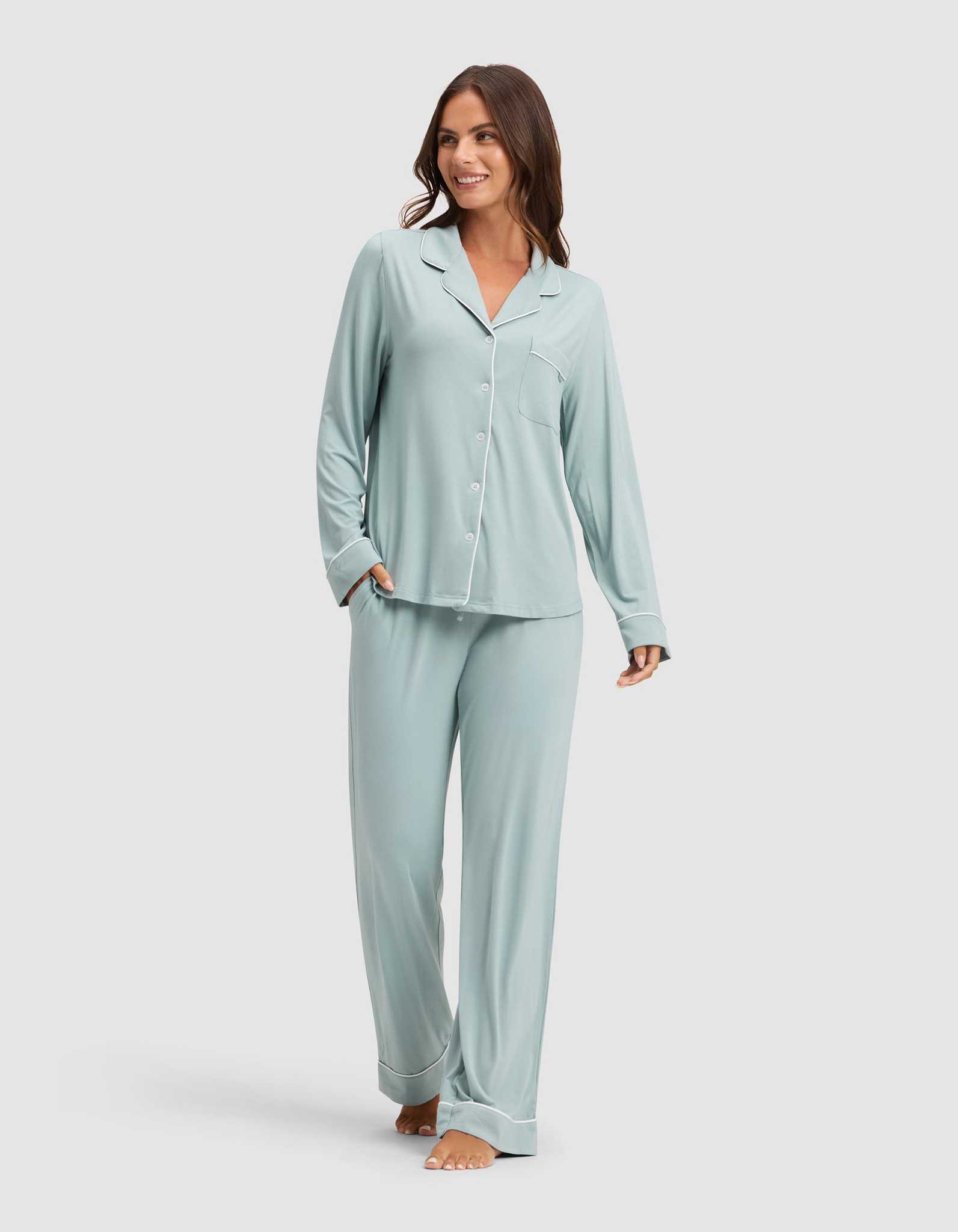 A woman smiles while wearing HIDE's Women's Bamboo Stretch-Knit Pajama Pant in light blue with a matching long-sleeve button-up top, barefoot against a plain light background. |Color:Brook