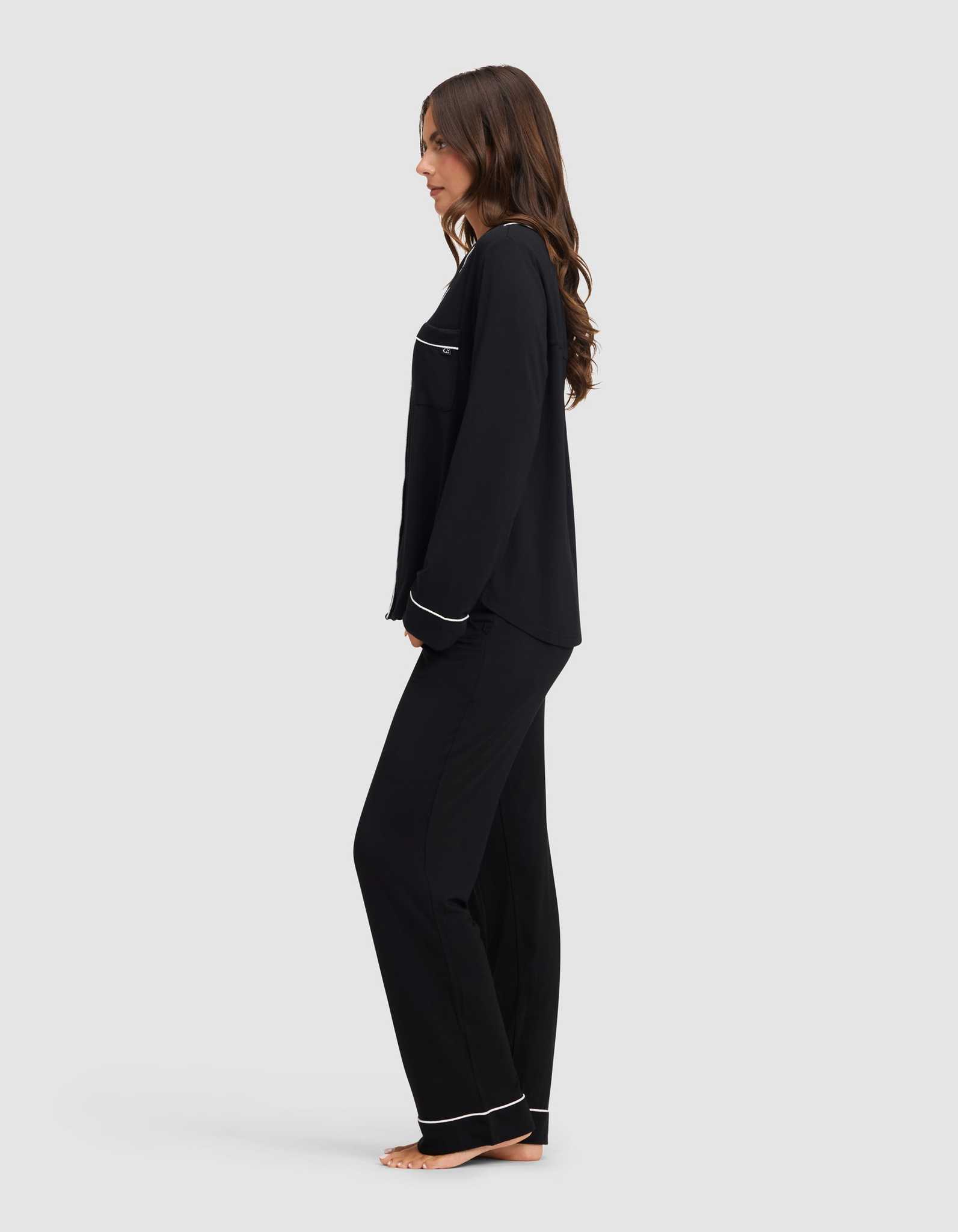 A woman stands barefoot in profile against a light gray background, wearing the Cozy Earth Women's Bamboo Stretch-Knit Long Sleeve Pajama Set in black with white trim. |Color:Black