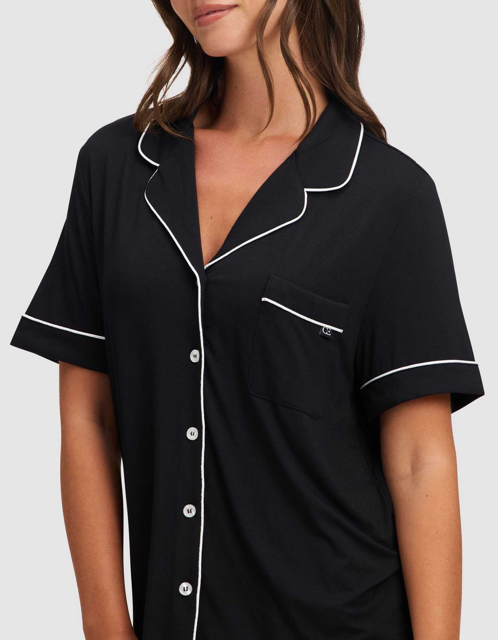 A woman wears the Cozy Earth Women's Bamboo Stretch-Knit Short Sleeve Pajama Set in black, featuring white piping, front buttons, and a chest pocket, standing against a plain light background. |Color:Black