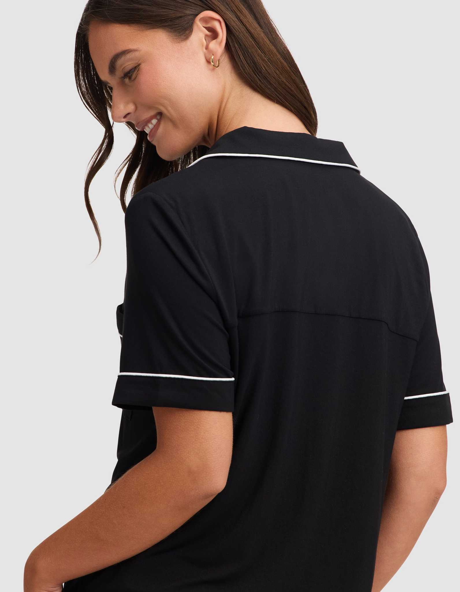 A woman with long brown hair smiles while looking down, wearing the HIDE Women's Bamboo Stretch-Knit Short Sleeve Pajama Top in black with white piping, shown from the back and side against a plain light gray background. |Color:Black