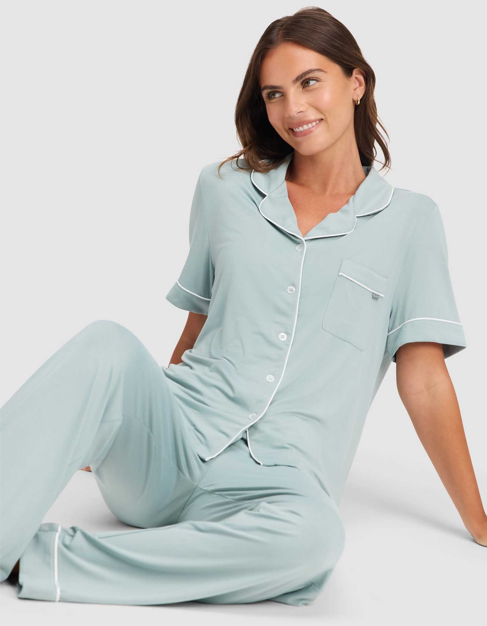 A woman with brown hair sits on the floor, smiling. She wears the HIDE Women's Bamboo Stretch-Knit Short Sleeve Pajama Top in light blue with white trim and matching long pants. The background is plain and light-colored. |Color:Brook