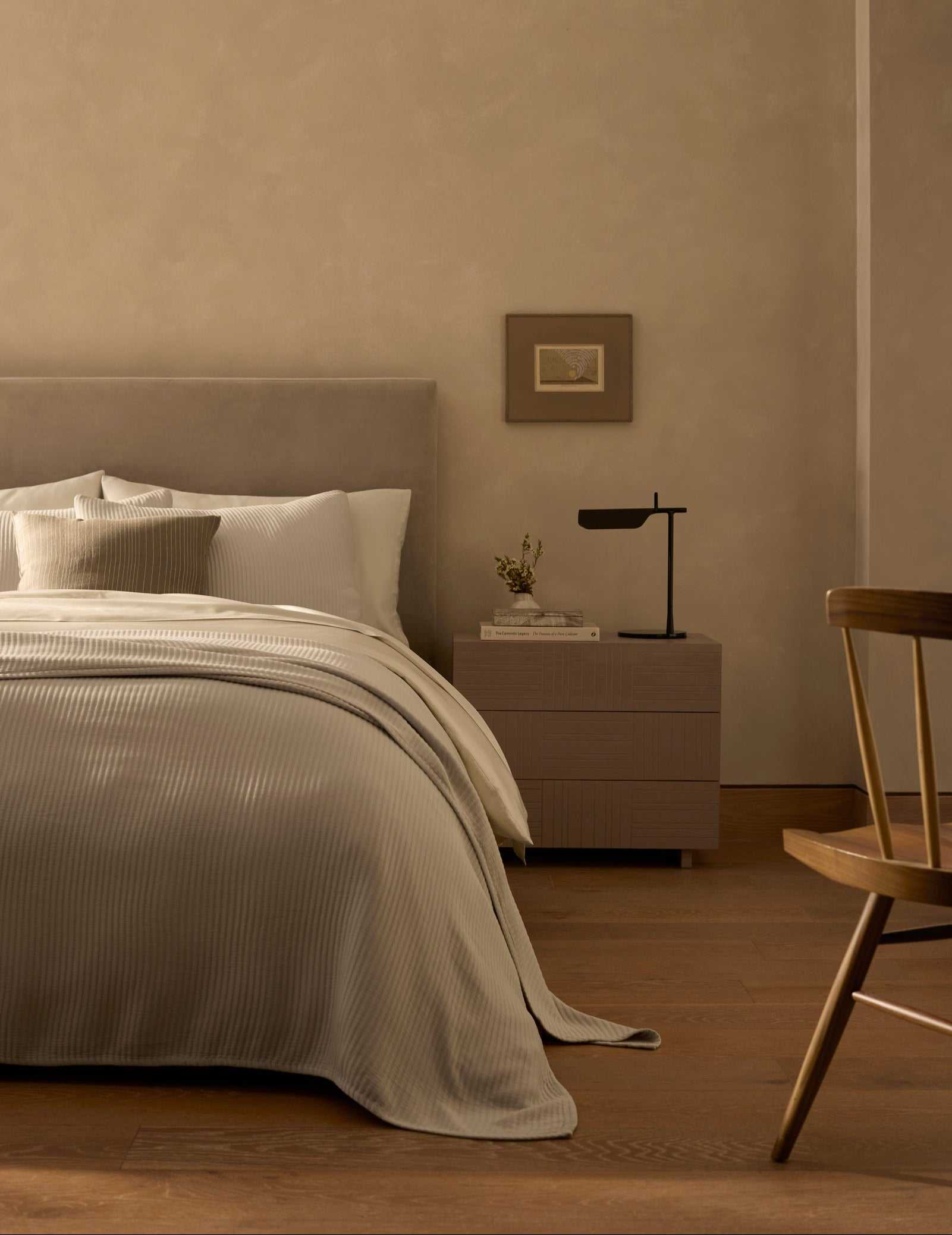 A minimalist bedroom features a neatly made bed with the Cozy Earth Bamboo Coverlet in beige tones, a wooden nightstand topped with a modern black lamp and small vase, a framed picture on the wall, and a wooden chair on hardwood floors.|Color:Driftwood