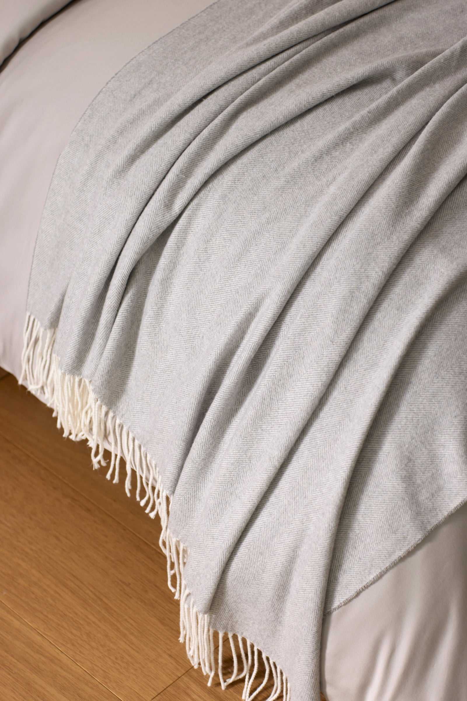 The Cozy Earth Bamboo Herringbone Tassel Throw in light gray is draped neatly over the edge of a bed with white bedding, set on a wooden floor.|Color:Cloud