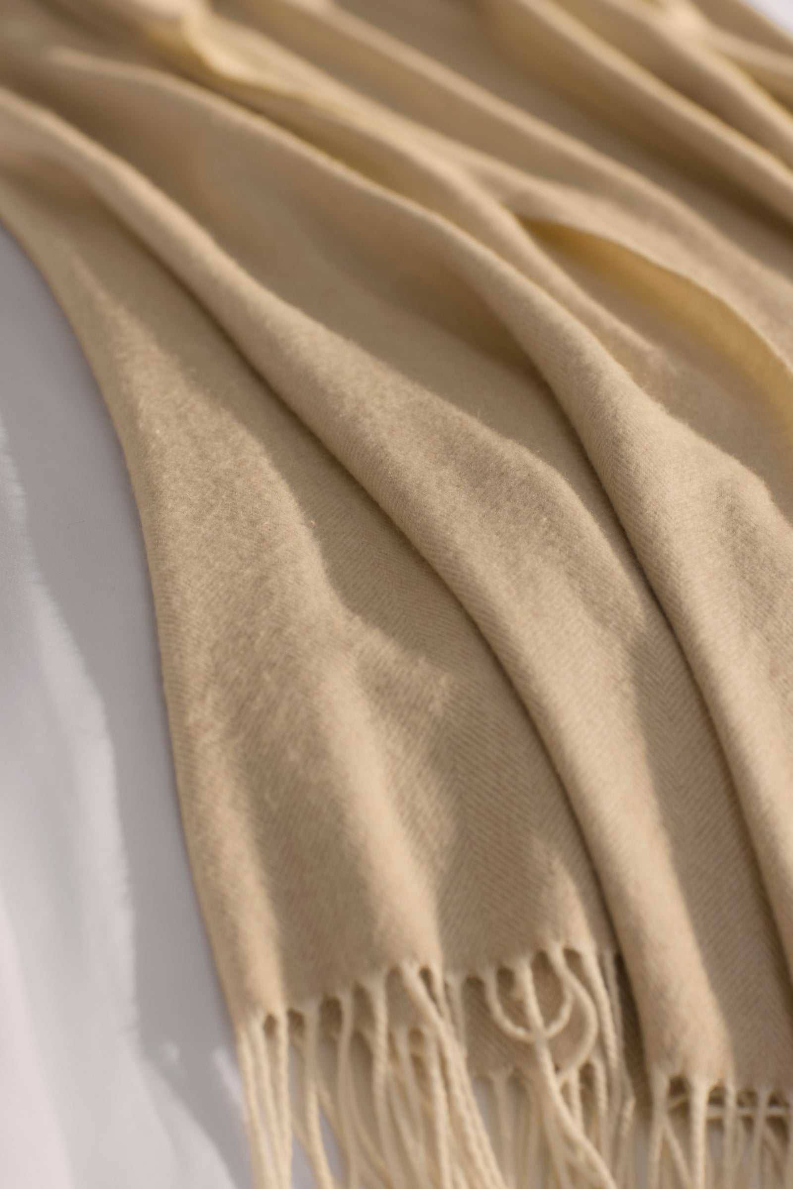 The Cozy Earth Bamboo Herringbone Tassel Throw, featuring a subtle herringbone texture and tasseled edges, is draped over a white surface in gentle folds.|Color:Oatmeal