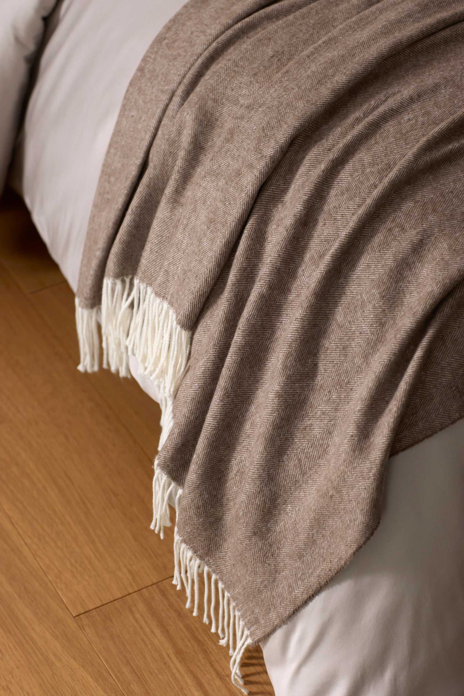 The Cozy Earth Bamboo Herringbone Tassel Throw, a soft light brown blanket with white fringe, is draped over a white bed resting on a wooden floor.|Color:Truffle