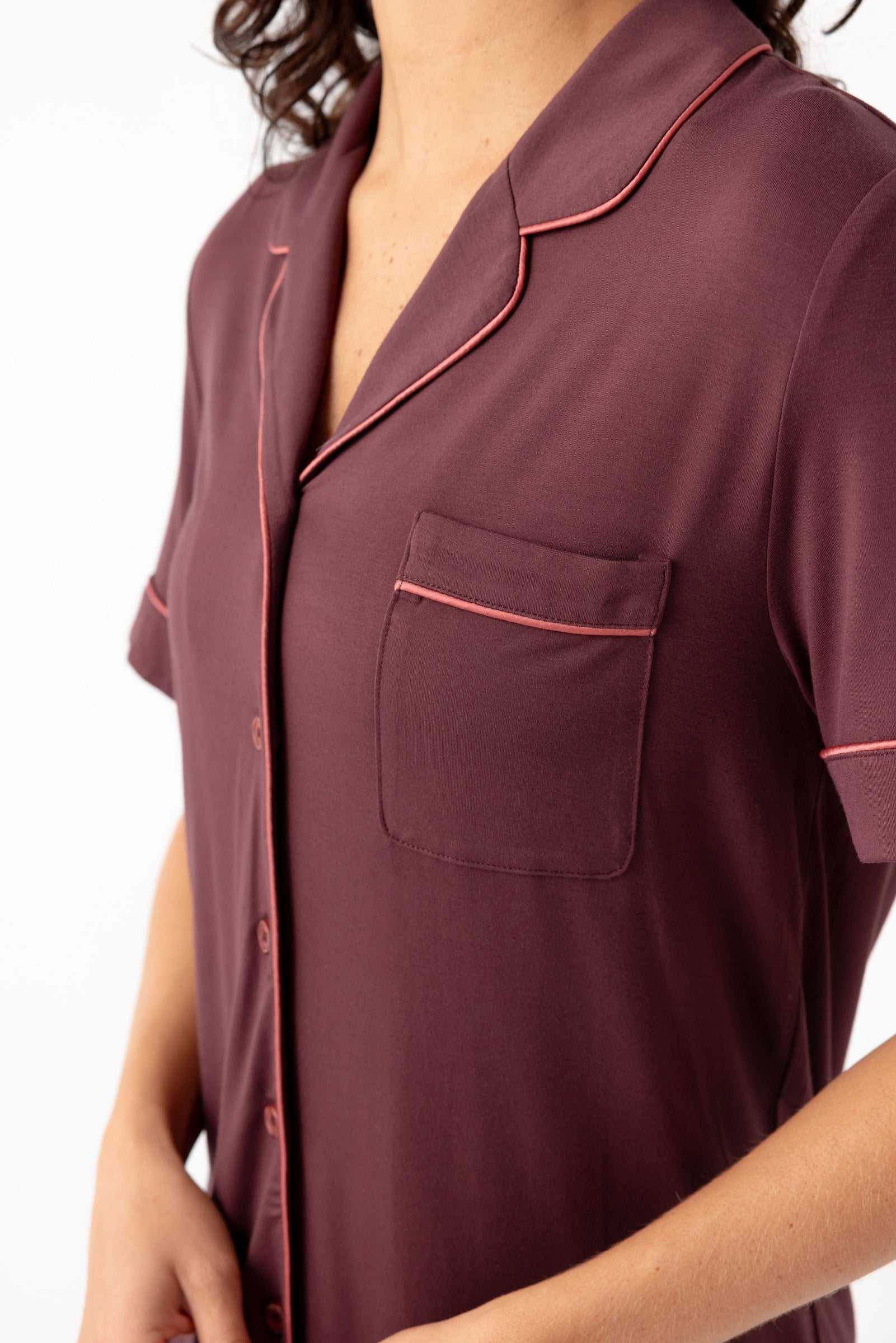 A person wears the Cozy Earth Women's Bamboo Stretch-Knit Short Sleeve Pajama Set—a maroon, pink-piped, button-down top with a breast pocket. The person's head is partially out of frame.|Color:Burgundy