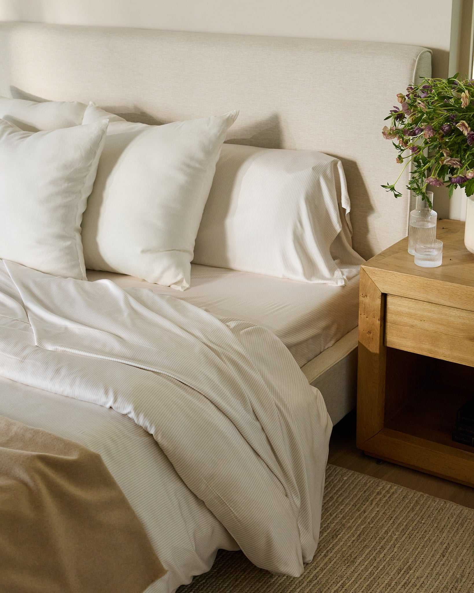 A cozy bedroom scene featuring a bed adorned with Cozy Earth's Bamboo Duvet Cover, complemented by white pillows and sheets, a beige blanket, and a light beige headboard. A wooden nightstand holds a potted plant and a glass of water, with books neatly stacked below. A window with greenery is in the background. |Color:Driftwood Pinstripe