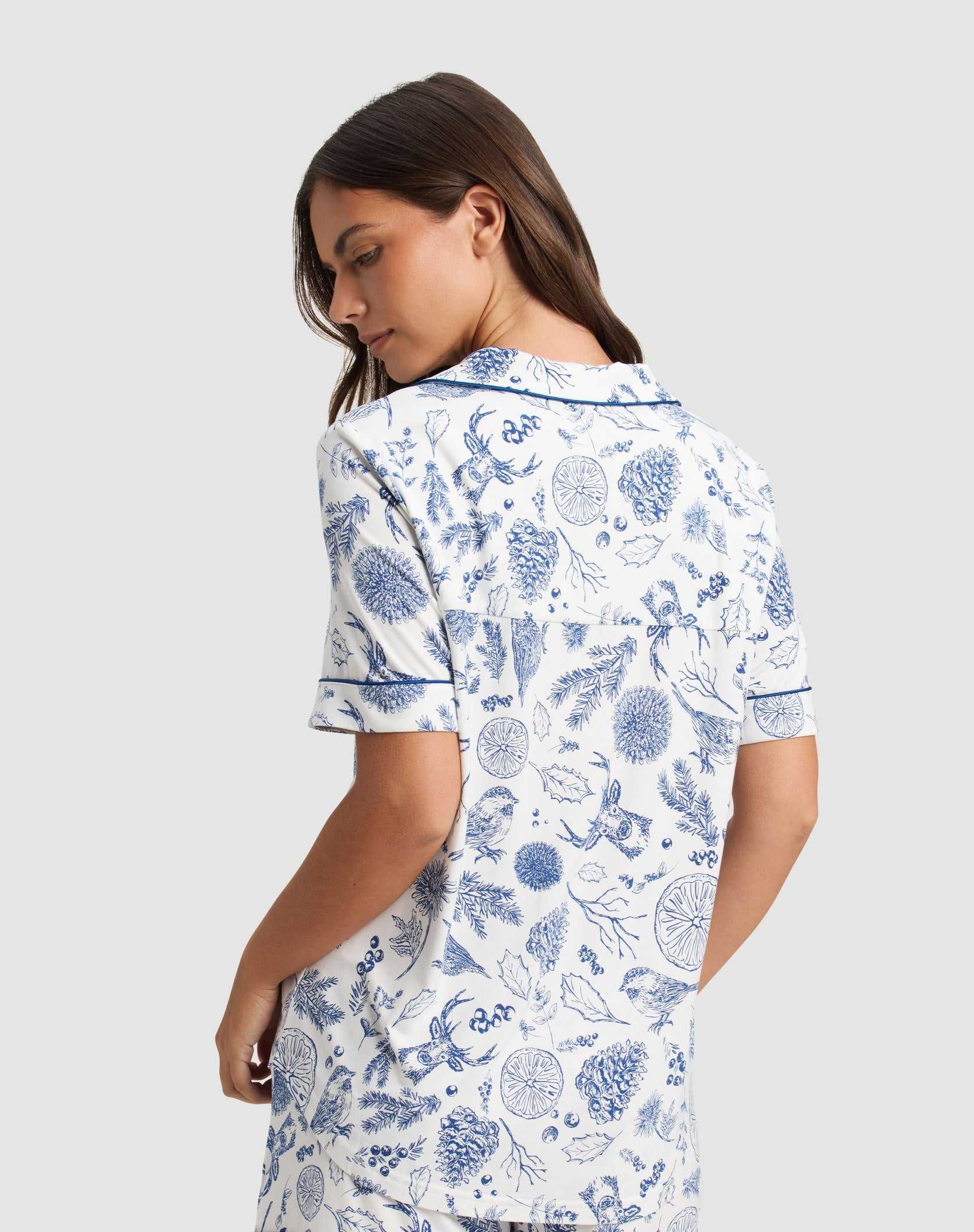 A woman stands turned slightly away, wearing the Cozy Earth Women's Bamboo Stretch-Knit Short Sleeve Pajama Top in white with blue botanical prints, a collar, and matching shorts. The background is plain light gray.|Color:Winter Toile Pacific