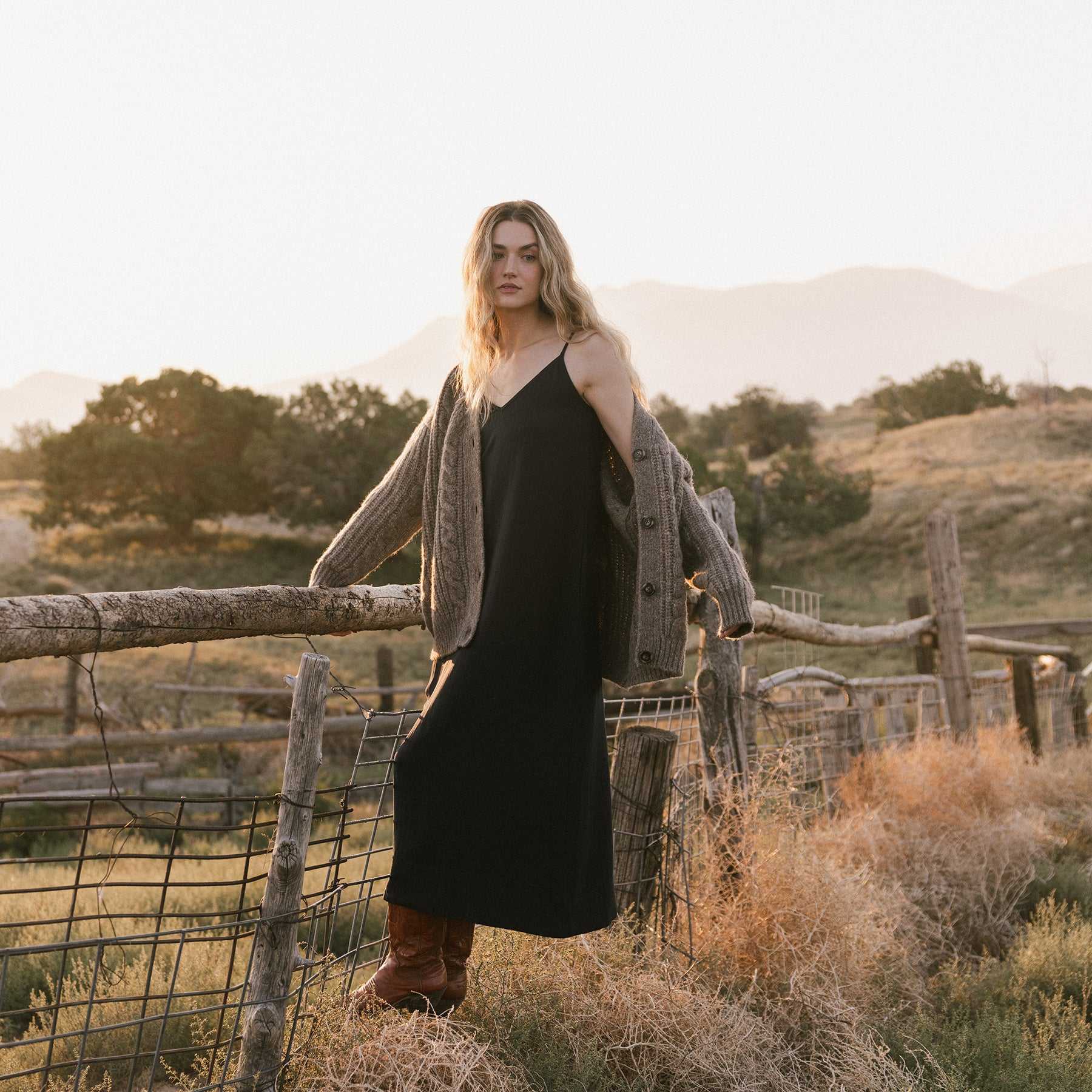 A person with long hair stands on a rustic fence in a rural setting, wearing a long black dress, an Oversized Cable Knit Cardigan by Cozy Earth, and brown boots. The background features rolling hills and sparse trees under a softly lit sky. |Color:Greige