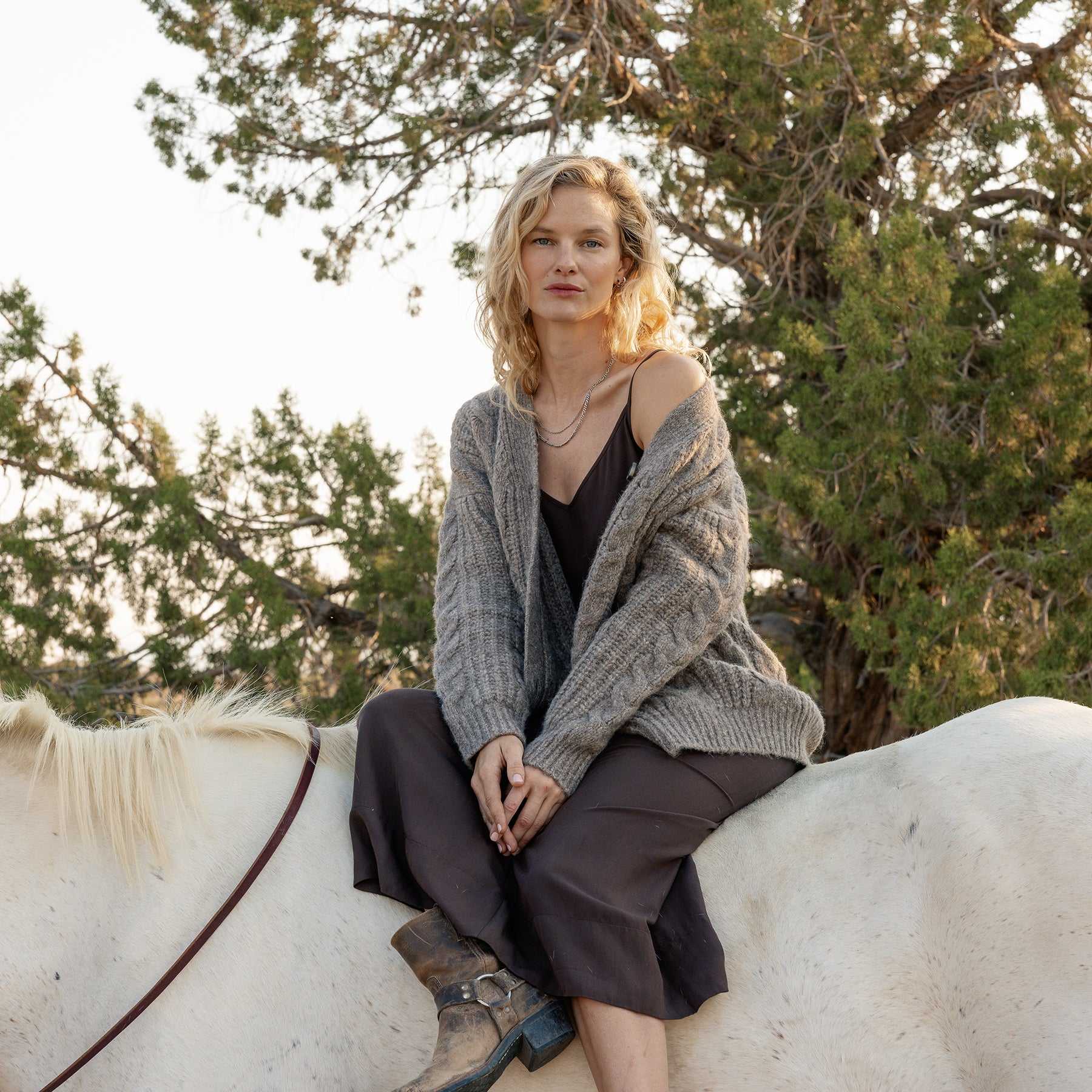 A person with blonde hair sits on a white horse, dressed in a brown dress and an Oversized Cable Knit Cardigan by Cozy Earth. They are surrounded by green trees in a natural setting, creating a calm and serene atmosphere. |Color:Oxford
