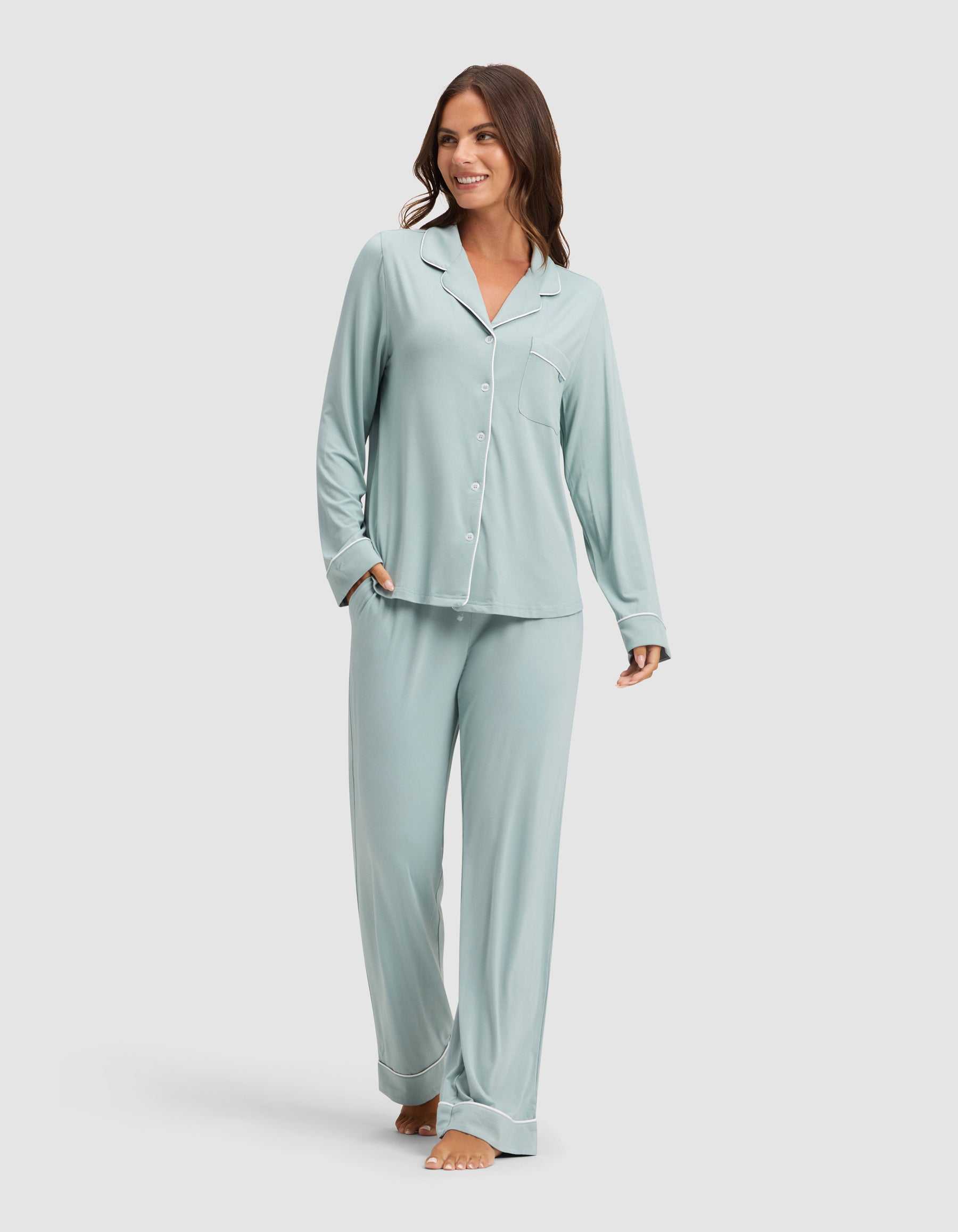 A woman smiles while wearing HIDE's Women's Bamboo Stretch-Knit Pajama Pant in light blue with a matching long-sleeve button-up top, barefoot against a plain light background. |Color:Brook