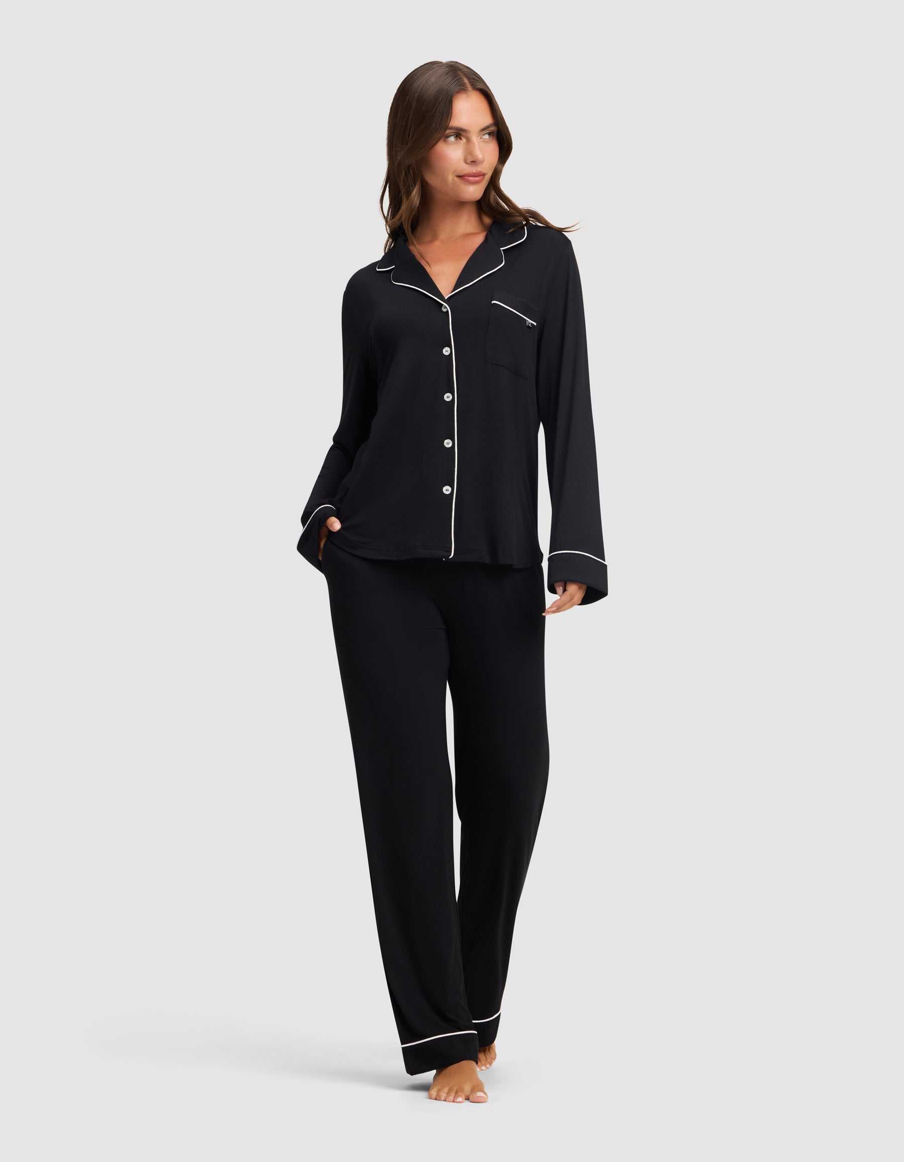 A woman stands barefoot against a light gray background, wearing Cozy Earth's Women's Bamboo Stretch-Knit Long Sleeve Pajama Set in black with white piping, featuring a button-up top and matching pants. |Color:Black