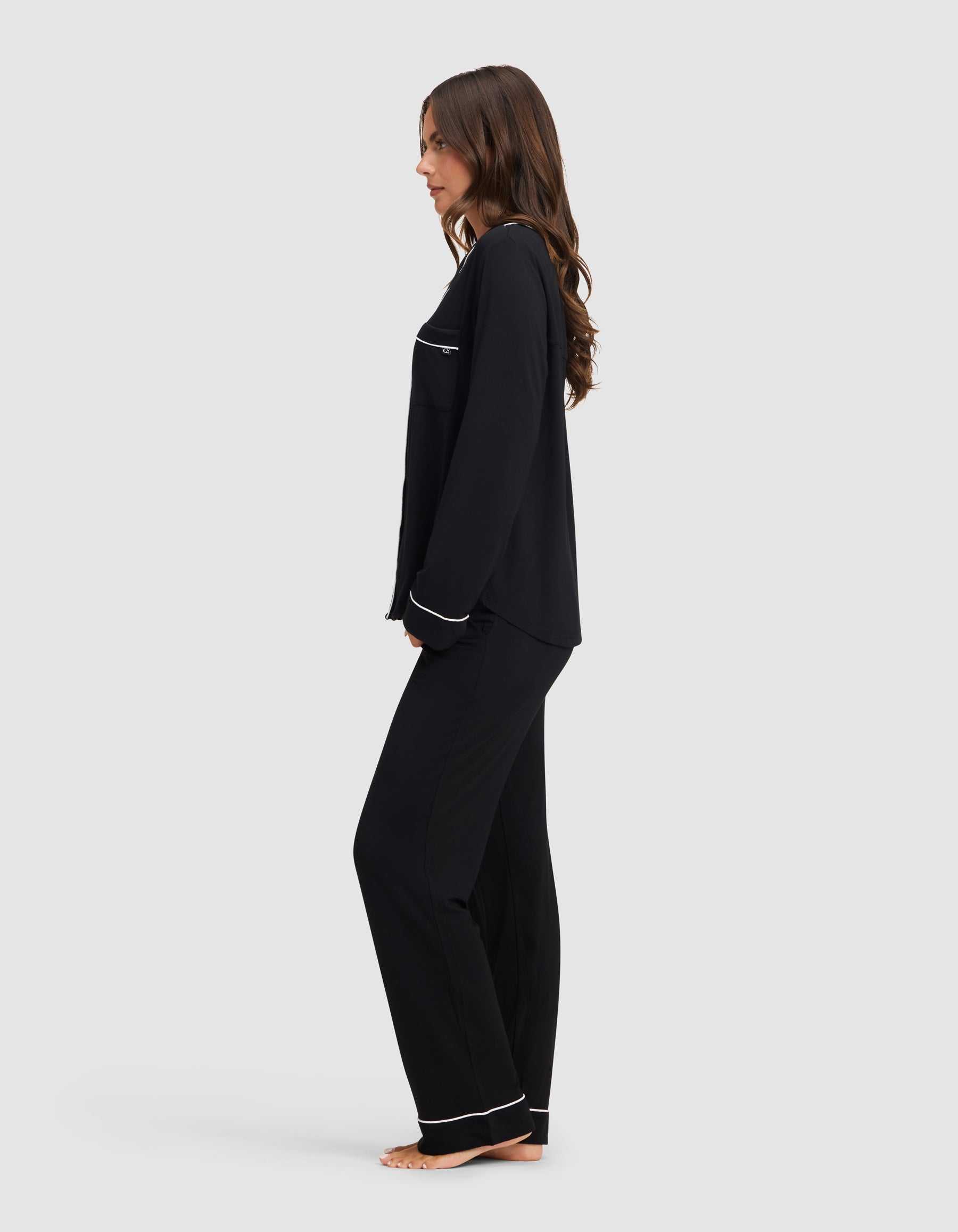 A woman stands barefoot in profile against a light gray background, wearing the Cozy Earth Women's Bamboo Stretch-Knit Long Sleeve Pajama Set in black with white trim. |Color:Black