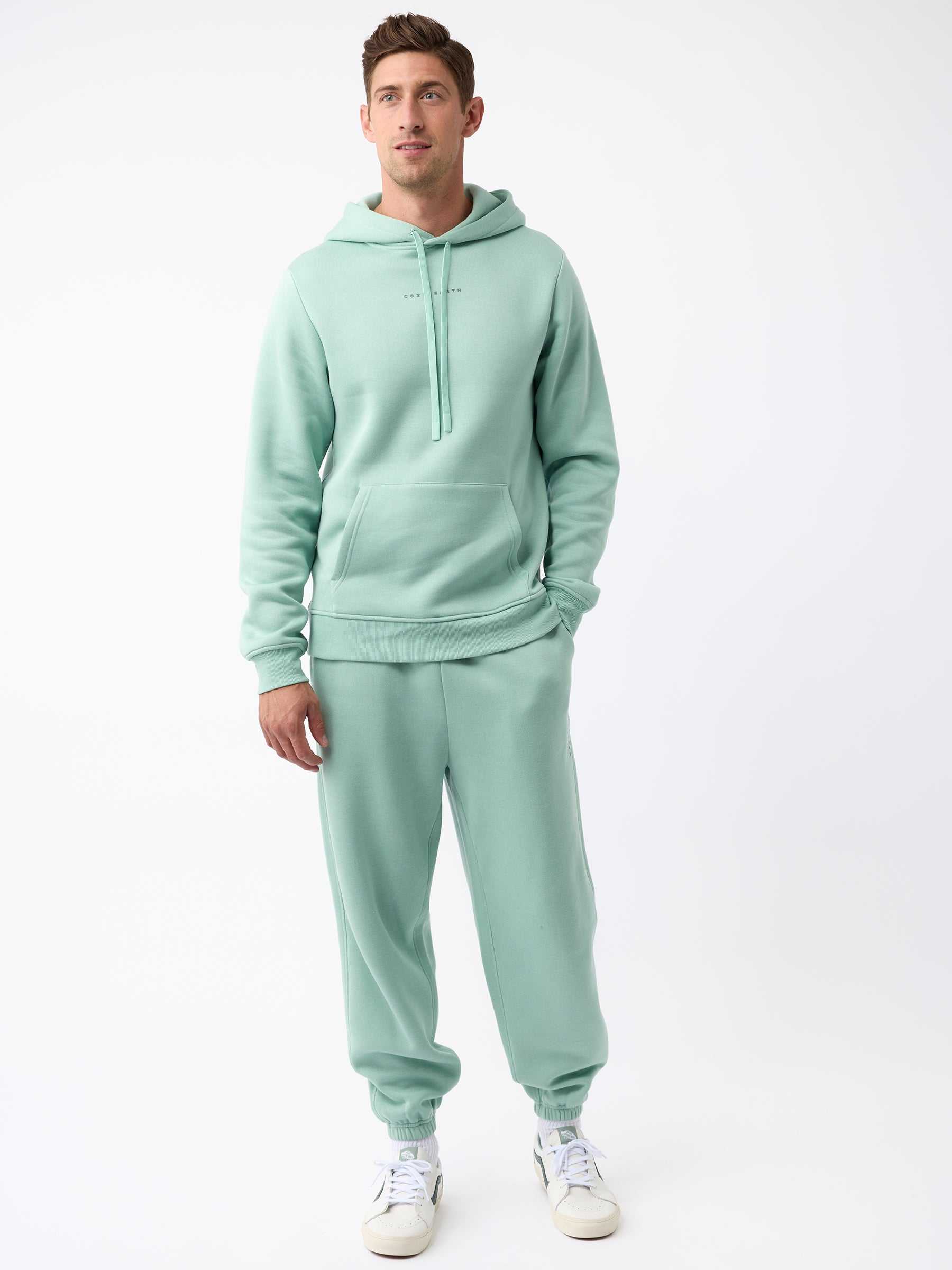 A man dressed in Cozy Earth's Men's CityScape Sweatpant in a light green hue stands against a plain white background. He completes his look with matching attire and white sneakers, his hands resting comfortably at his sides as he gazes slightly to the left. |Color:Wintergreen