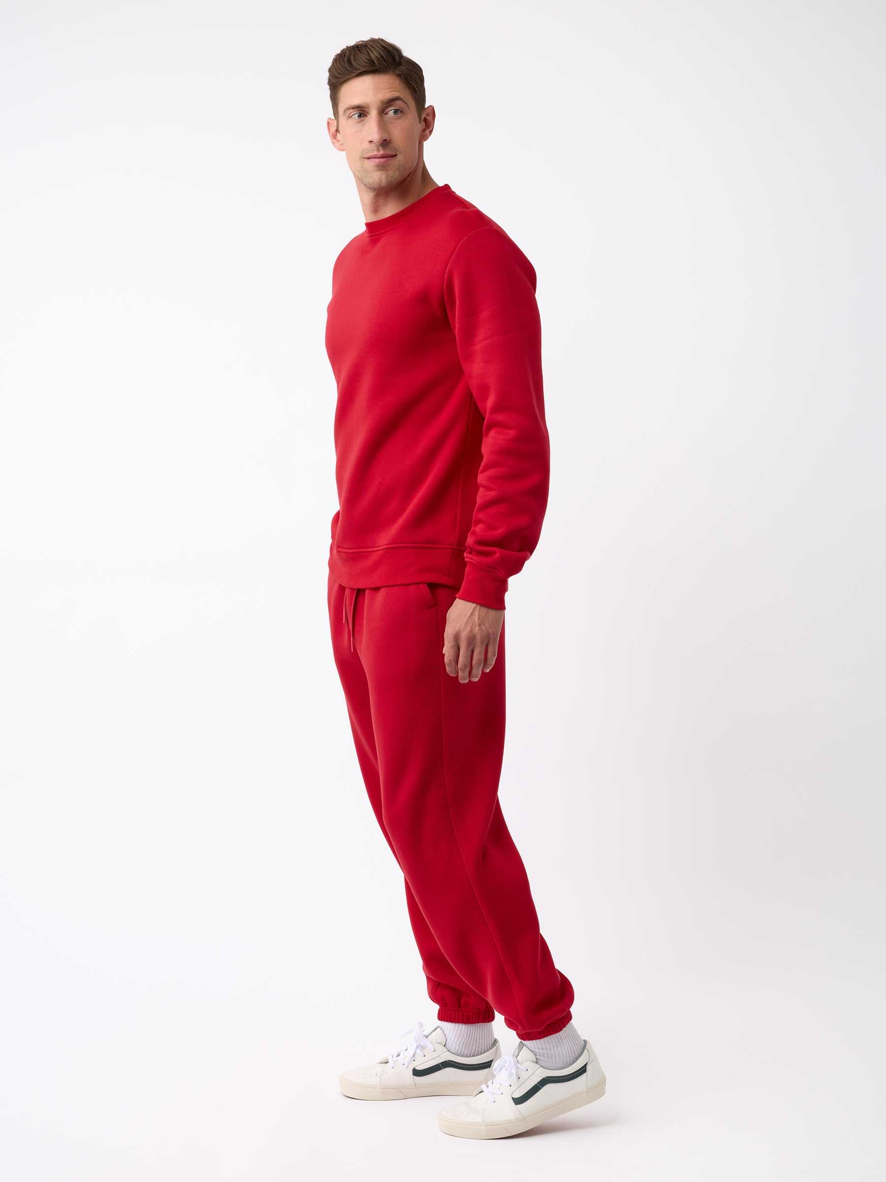A man dressed in a red CityScape Crewneck by Cozy Earth stands against a white background, facing sideways. He is wearing white sneakers and looking slightly to the left. |Color:Crimson