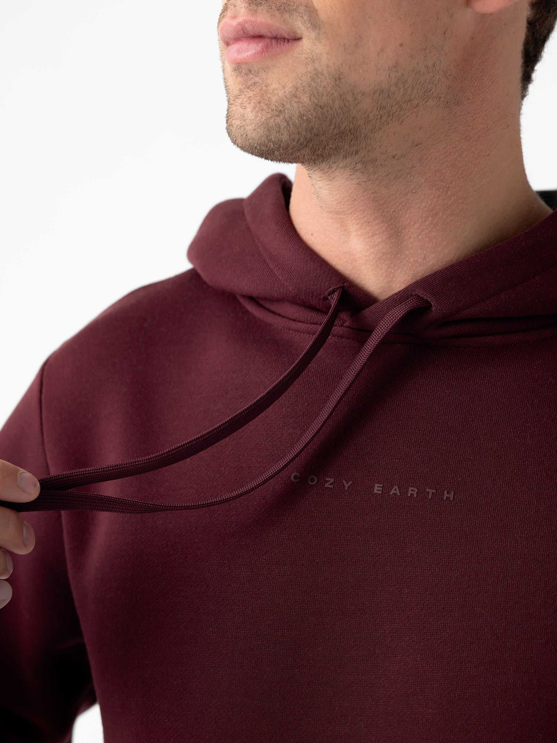 Close-up of a person wearing a maroon Men's CityScape Hoodie with the text "Cozy Earth" written on it, lightly tugging at one of the drawstrings. The face is partially out of frame, and the background is plain white. |Color:Burgundy