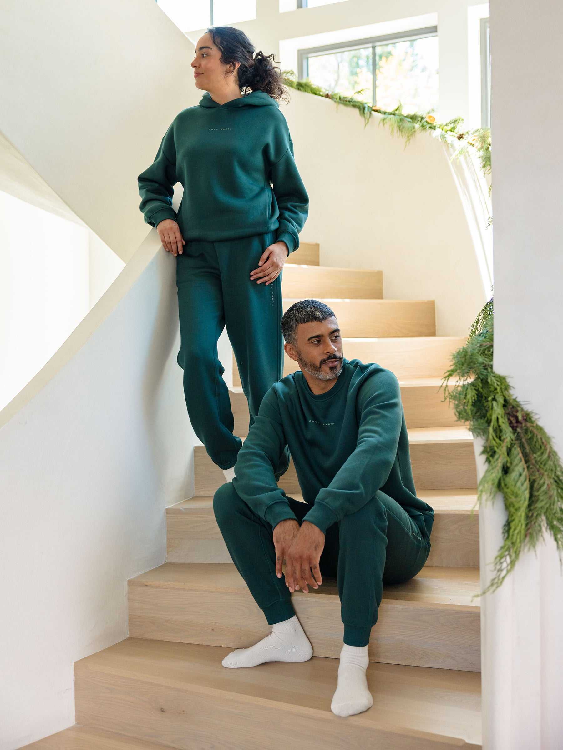 Two individuals, both donning the Men's CityScape Jogger by Cozy Earth in matching green, unwind on wooden stairs decorated with lush greenery. One person is seated on a step while the other stands with one foot on the step, gazing into the distance in a sunlit room. |Color:Spruce