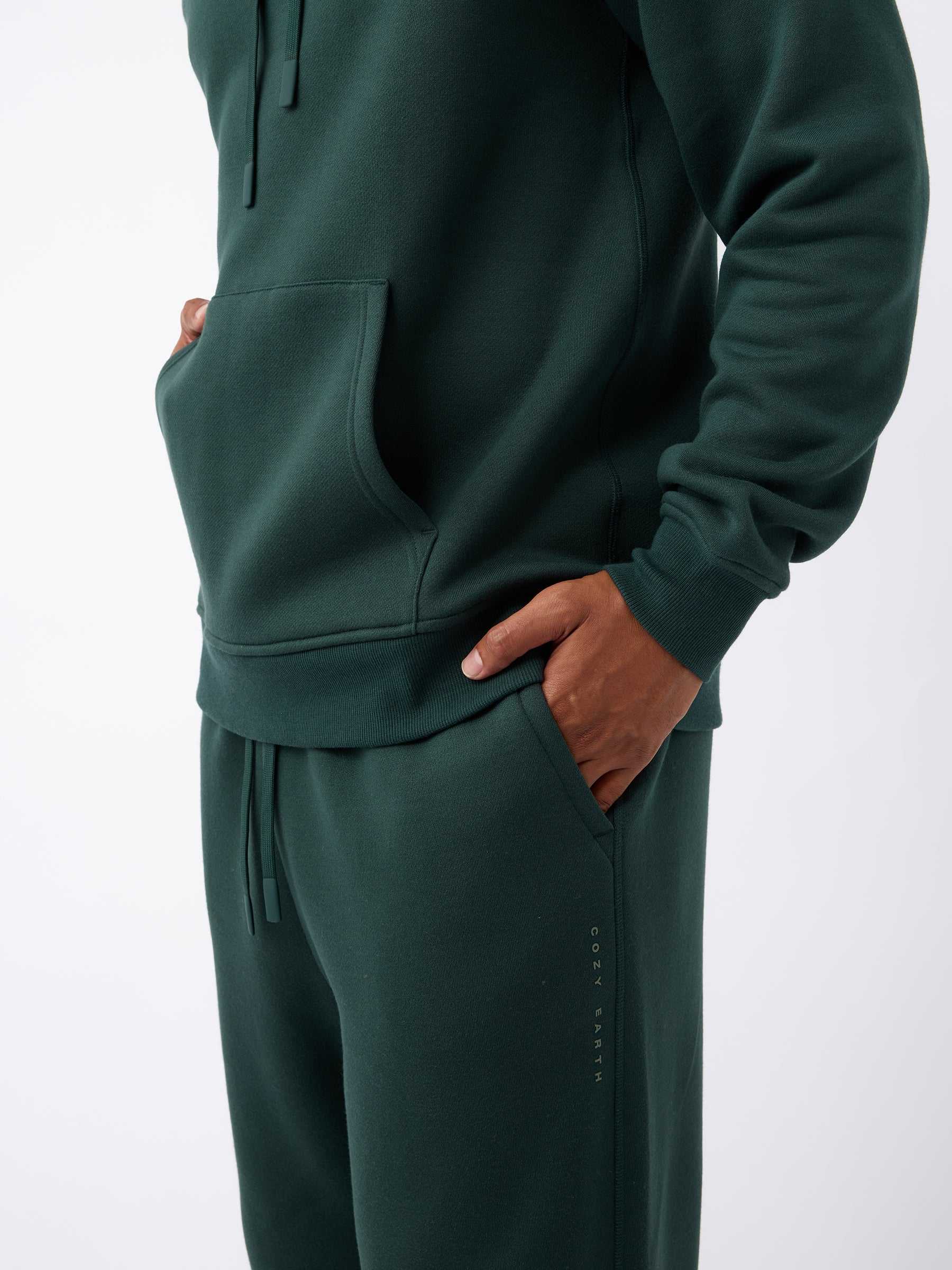 A person is wearing a dark green hoodie and matching Men's CityScape Sweatpants from Cozy Earth, with hands in pockets. The clothing features a subtle logo that reads "COZY EARTH" on the pants, set against a plain white background. |Color:Spruce