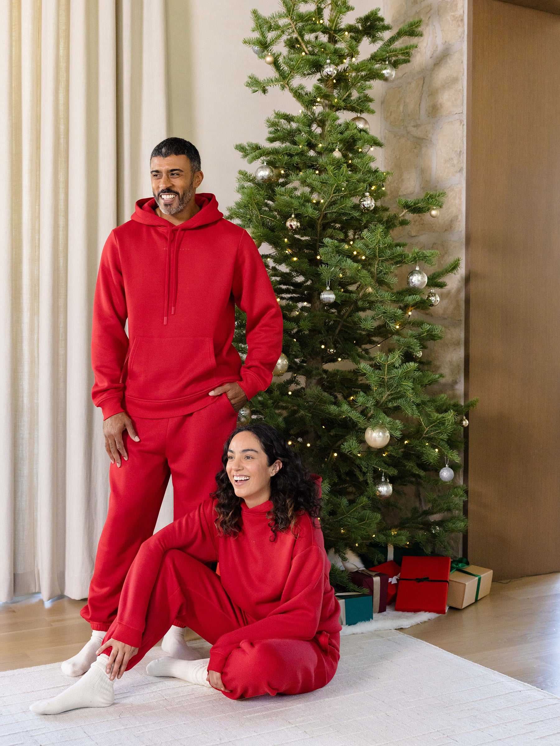 A man and a woman wearing matching Men's CityScape Hoodies from Cozy Earth are in a cozy living room decorated for Christmas. The woman sits on the floor, smiling, while the man stands behind her. A Christmas tree adorned with ornaments and presents is visible in the background. |Color:Crimson