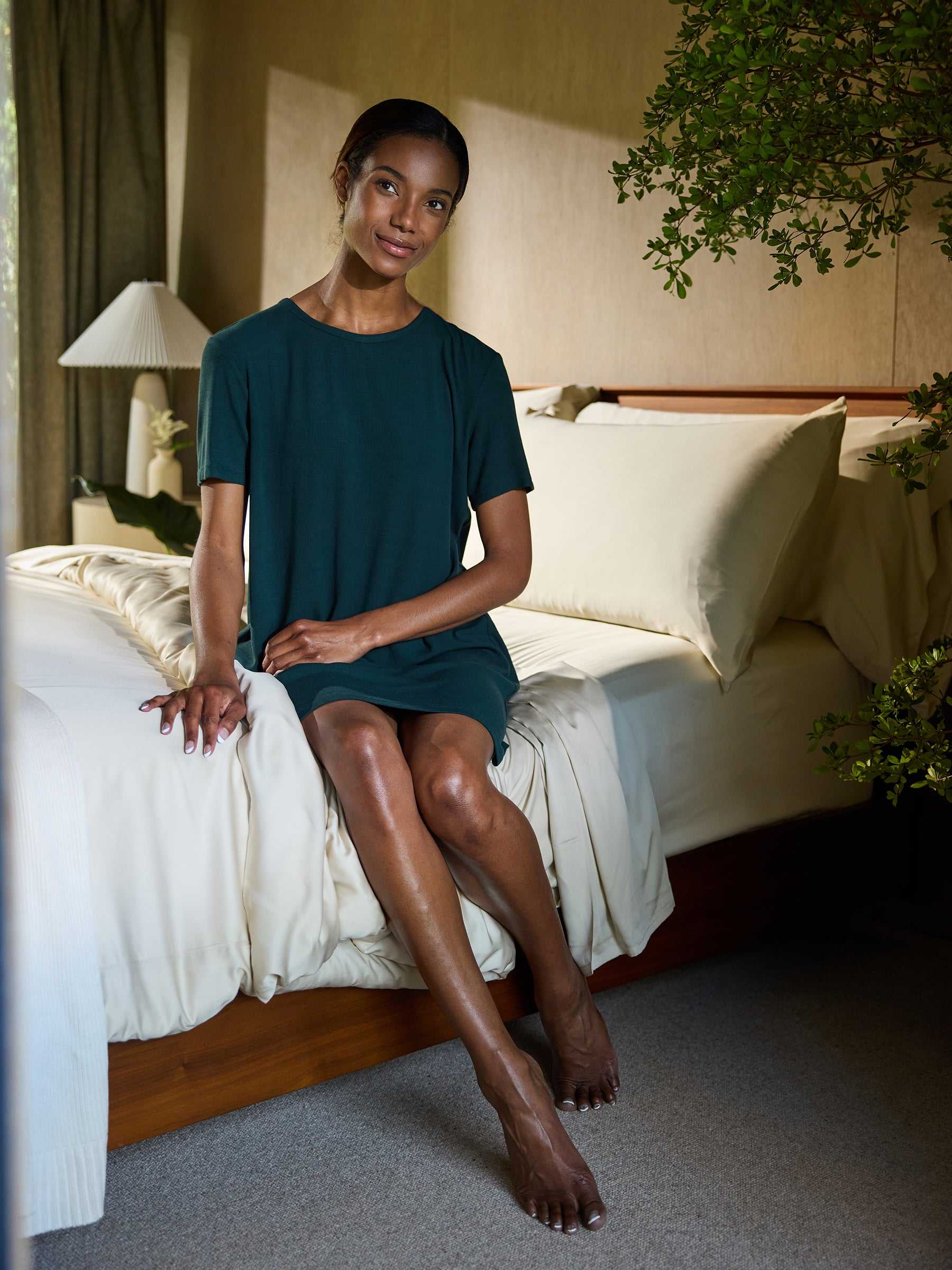 A woman wearing a Women's Bamboo Rib-Knit Boyfriend Sleep Dress by Cozy Earth sits on a bed with beige linens. She is resting her hands on the bed and looking at the camera with a slight smile. The bedroom is adorned with green indoor plants and has a cozy, well-lit ambiance. |Color:Spruce
