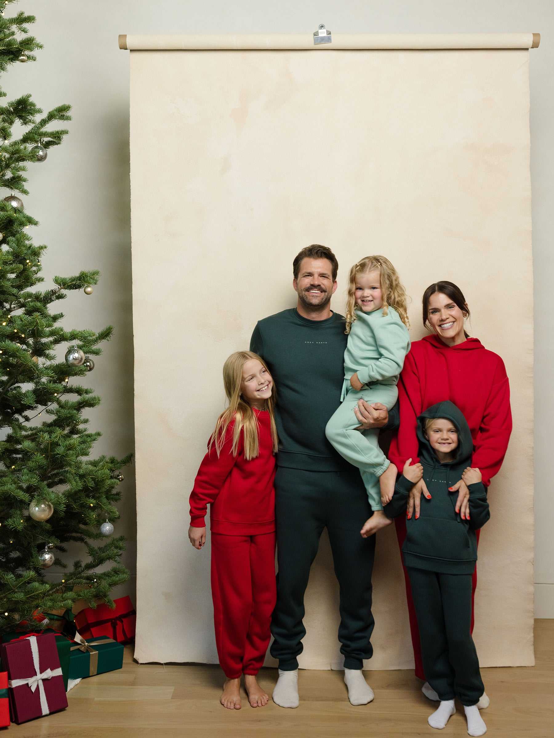 A family of five poses for a holiday photo with the father holding the youngest child. They are all dressed in festive red and green outfits from Cozy Earth. To the left, there's a decorated Christmas tree with presents, set against a beige backdrop. |Color:Spruce