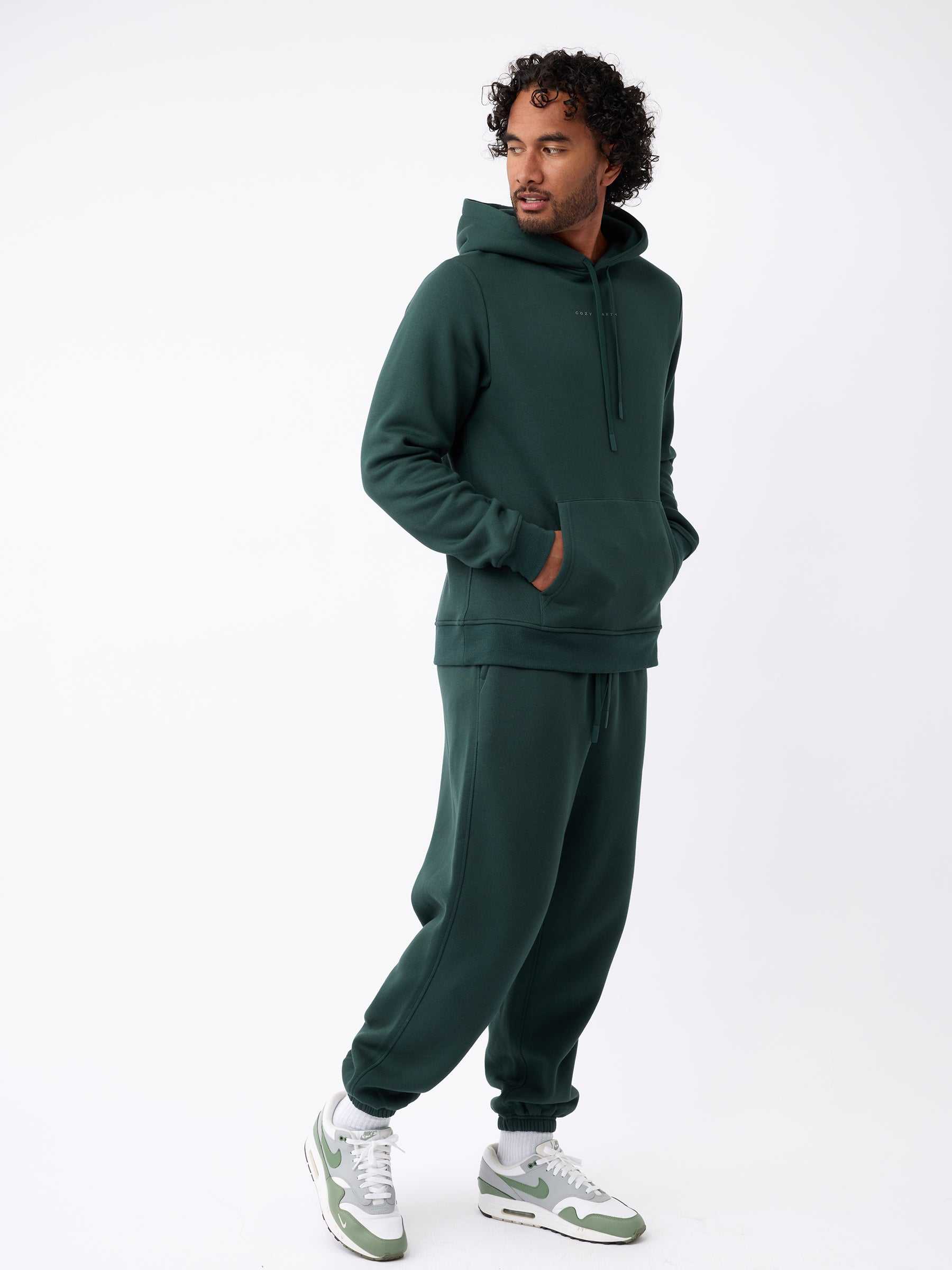 A man stands in a relaxed pose wearing a dark green hoodie paired with Cozy Earth's Men's CityScape Sweatpant. He has one hand in his pocket while looking to the side, and he complements his outfit with white and green sneakers against a white background. |Color:Spruce