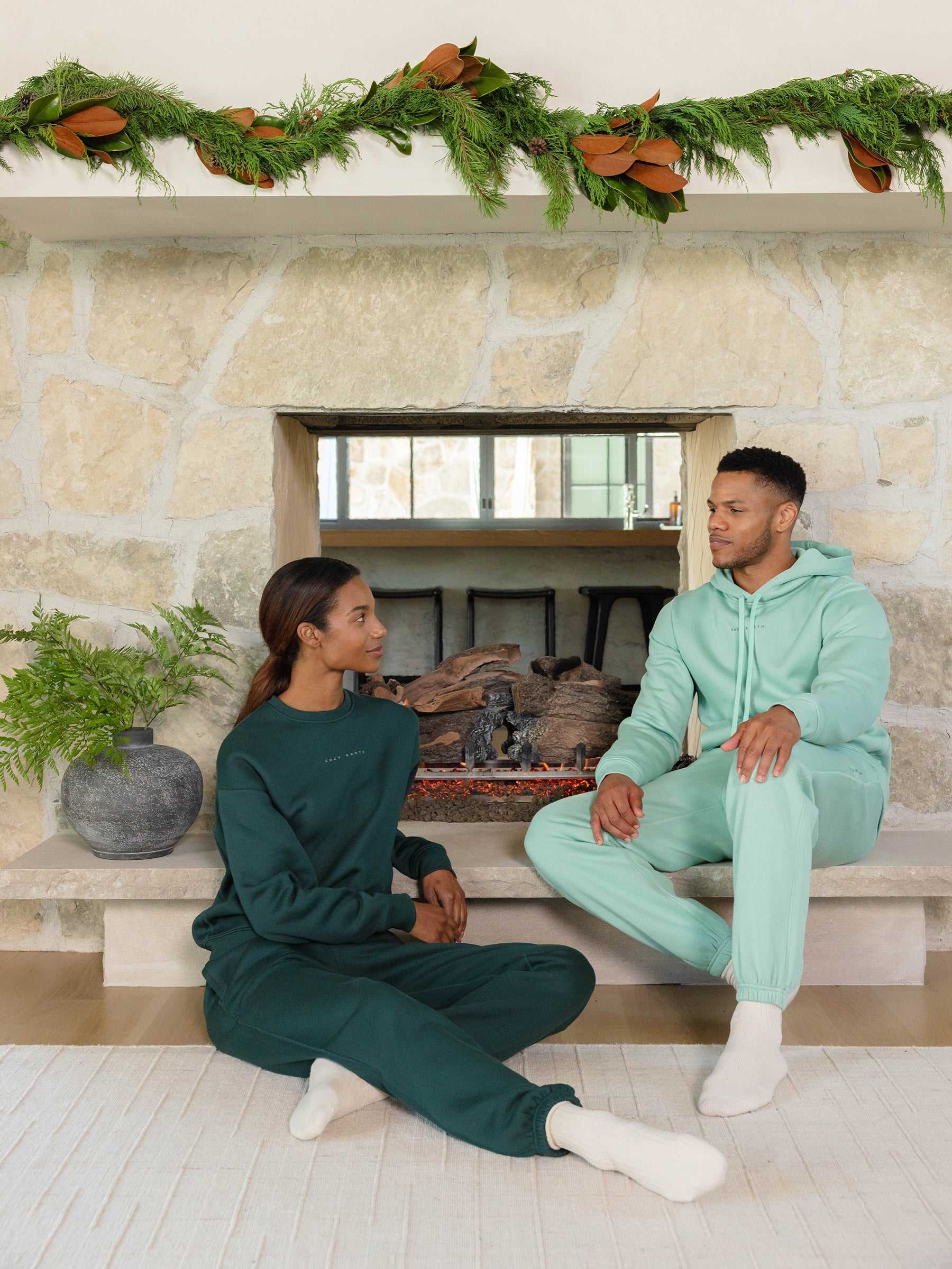 A man and woman dressed in matching green CityScape Hoodies from Cozy Earth sit together in front of a stone fireplace. Above the fireplace, a garland is draped, and a potted plant is placed nearby. They gaze at each other, enveloped in the room's cozy atmosphere. |Color:Wintergreen