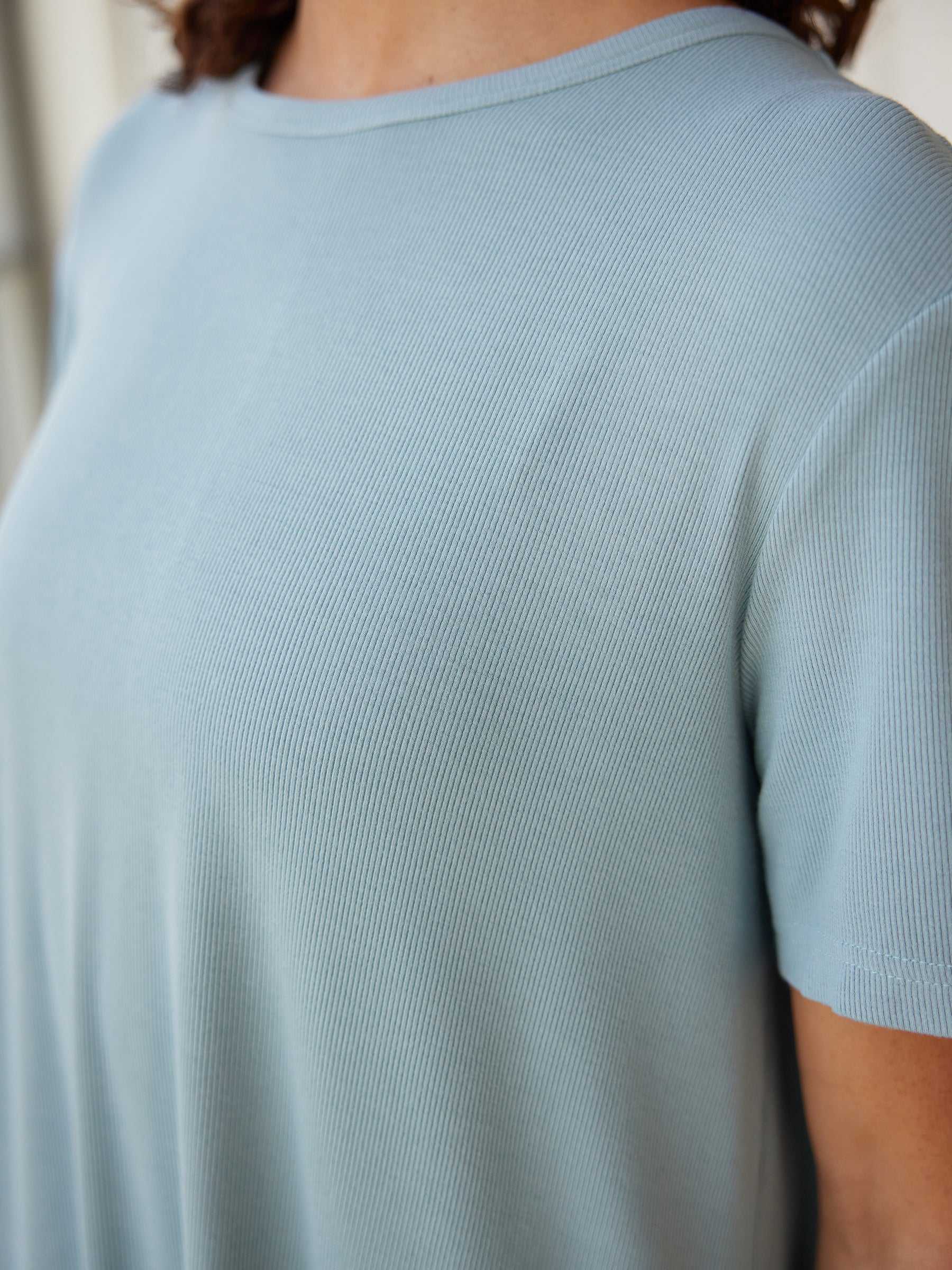 Close-up of a person wearing the Women’s Bamboo Rib-Knit Boyfriend Sleep Dress in light blue by Cozy Earth. The image focuses on the texture and fabric of the dress, showcasing detailed ribbing and soft material. The person's right shoulder and neck area are partially visible. |Color:Smokey Blue