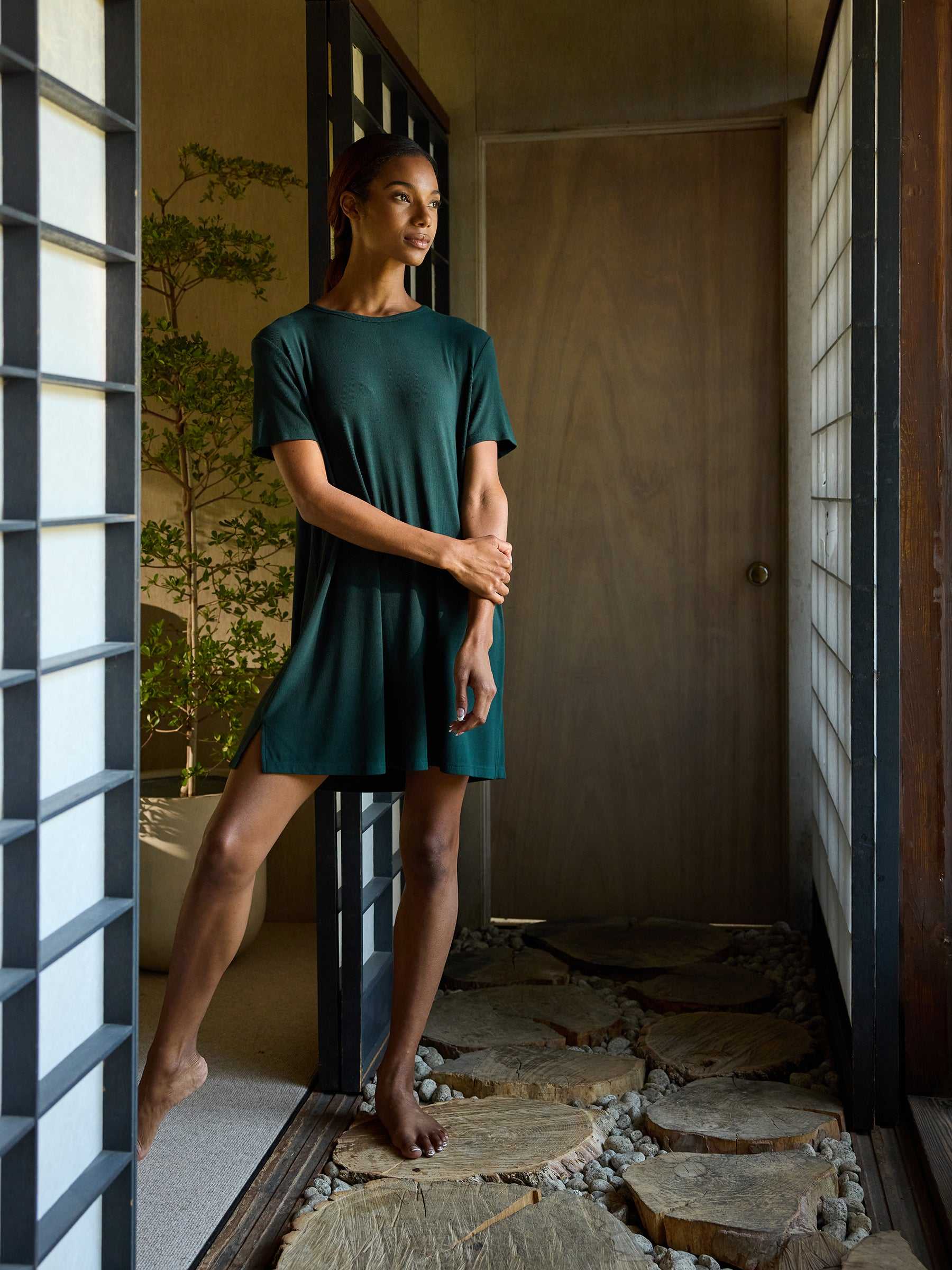 A woman in the Women’s Bamboo Rib-Knit Boyfriend Sleep Dress by Cozy Earth stands in a serene, minimalist hallway with wooden elements and natural light. She is barefoot, with one hand on her hip and the other gently resting on her arm. Potted plants and a stone floor add to the tranquil atmosphere. |Color:Spruce