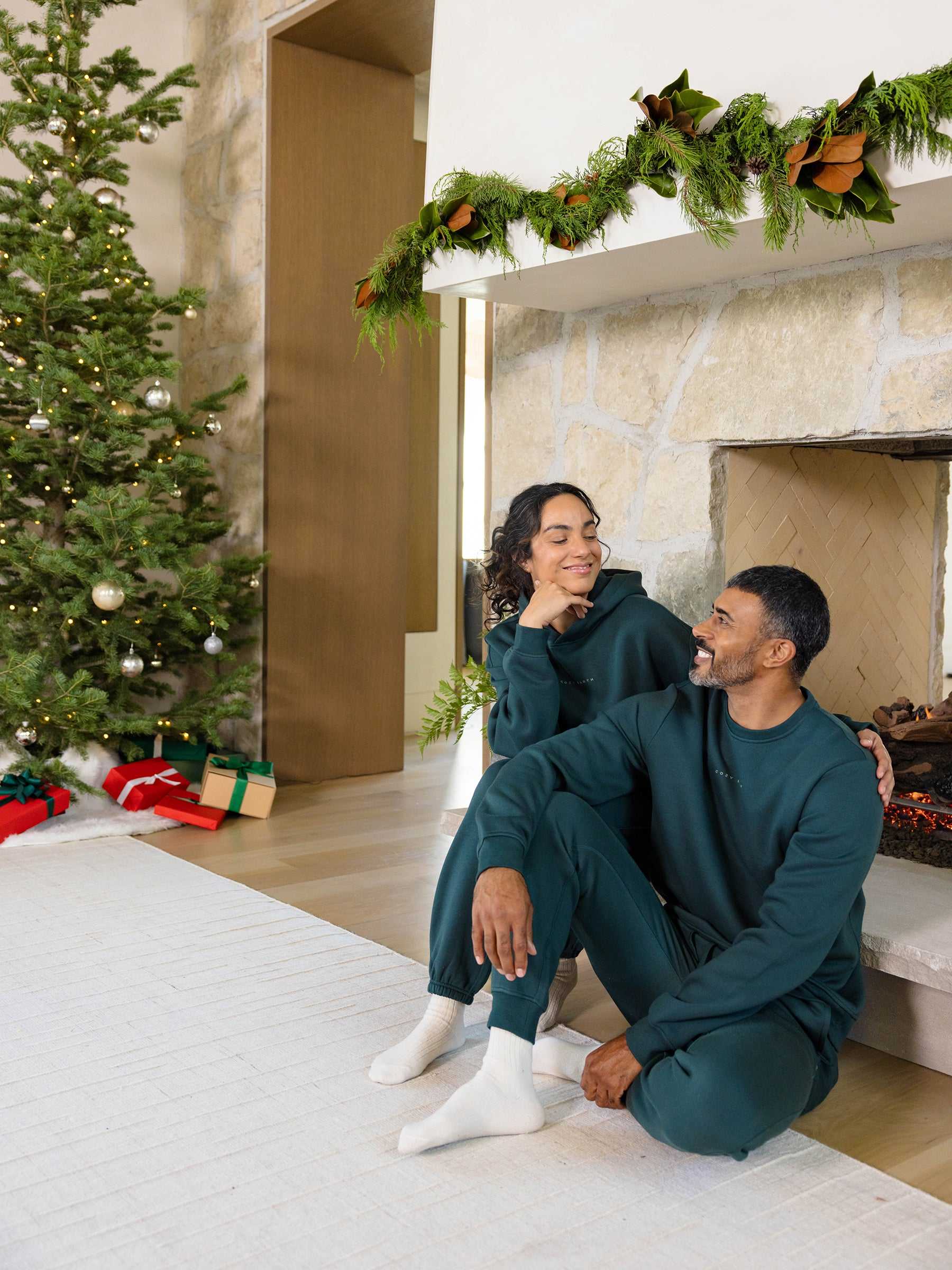Dressed in matching Men's CityScape Crewnecks by Cozy Earth, a couple sits by the fireplace with warm smiles. A beautifully decorated Christmas tree and an array of gifts are visible in the background, while the mantel boasts lush greenery, creating a cozy and festive holiday atmosphere. |Color:Spruce